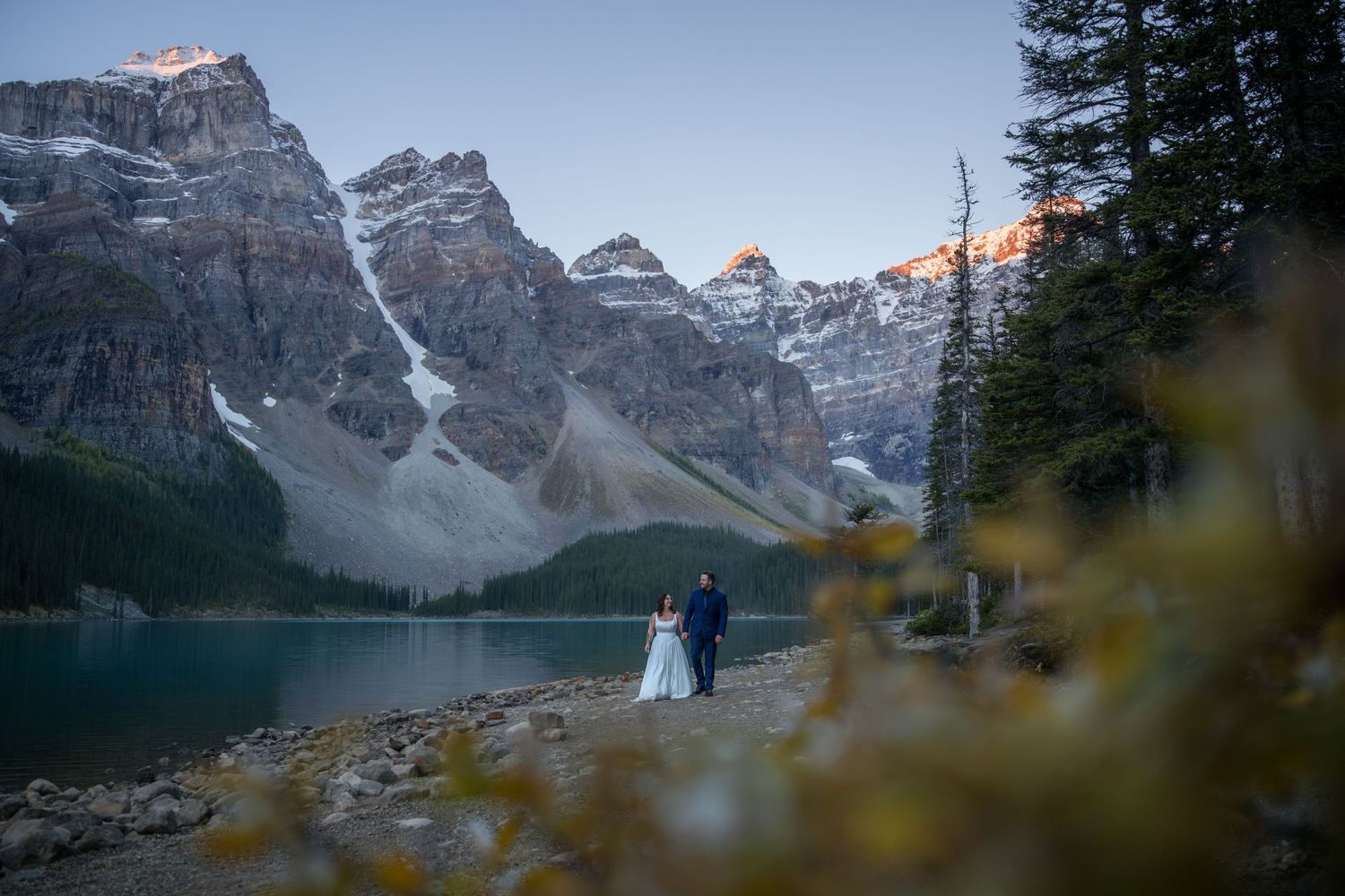Bride and groom stand by turquoise Moraine Lake with dramatic Rocky Mountain peaks at sunset in Banff National Park.