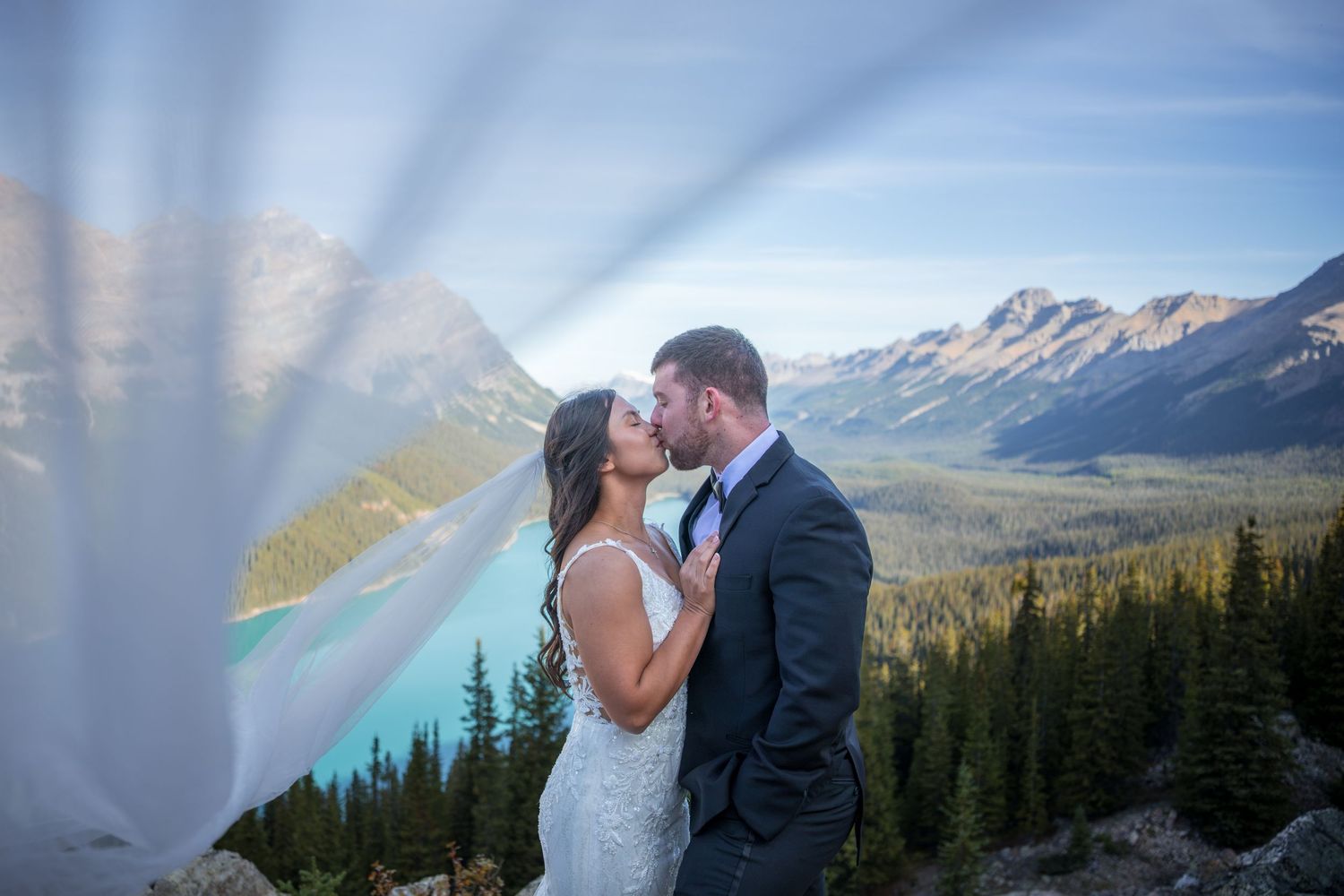 Bride and groom kissing with veil flowing, turquoise lake and mountain peaks in background at Peyto Lake.