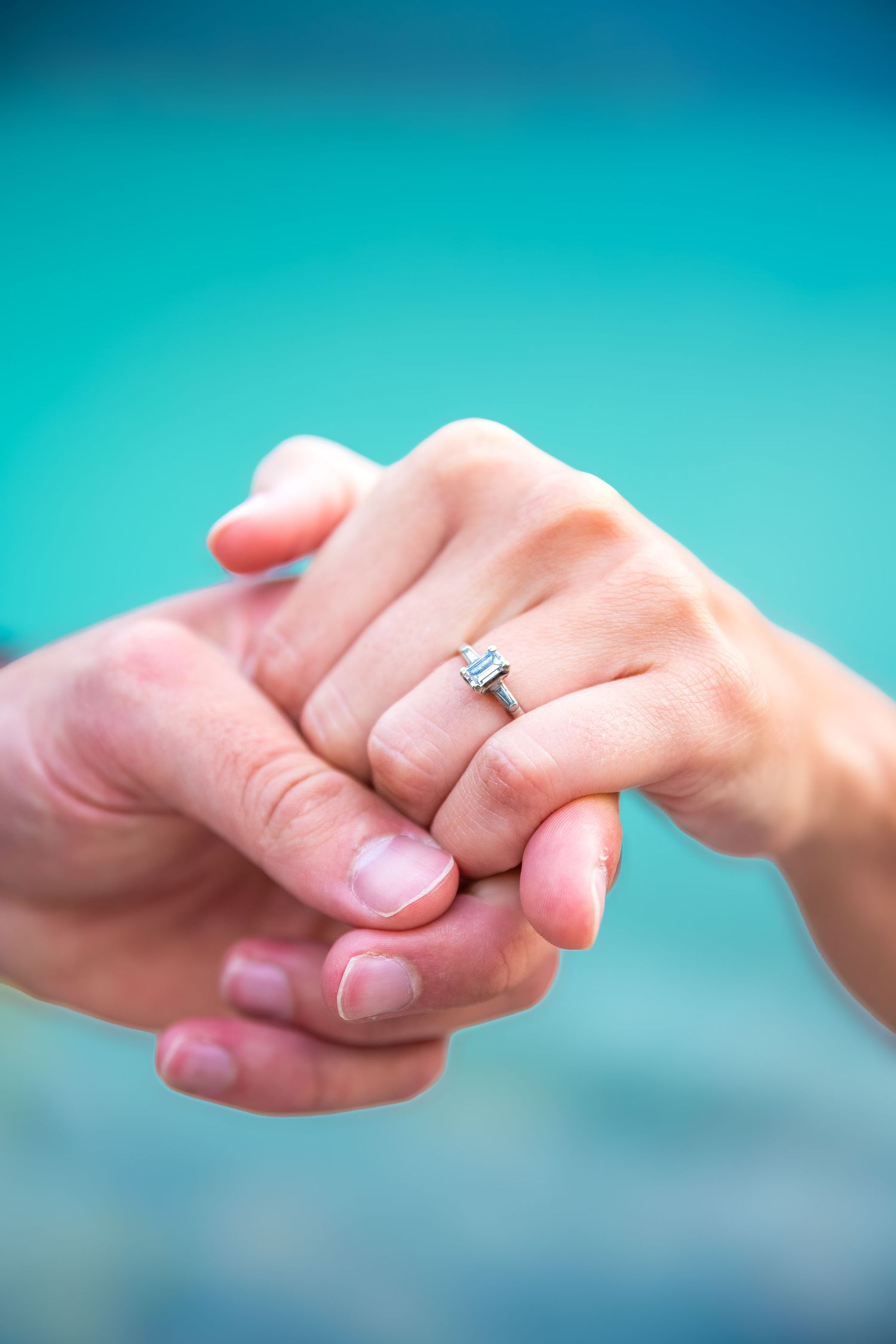 Two hands holding together with a sparkling diamond engagement ring on the woman's finger against a turquoise background.