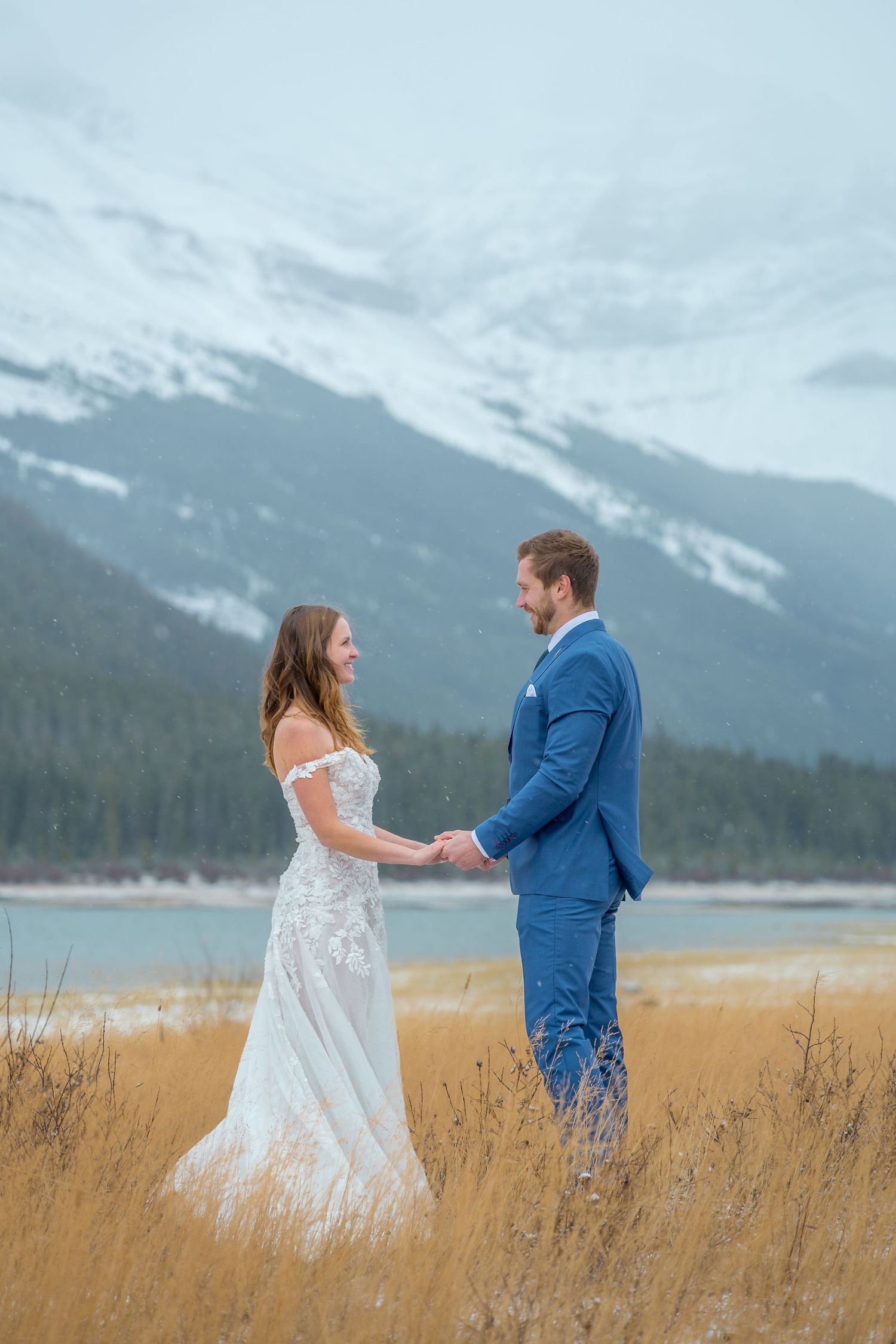 Bride and groom hold hands in a snowy mountain landscape with golden grass and a lake in the background.