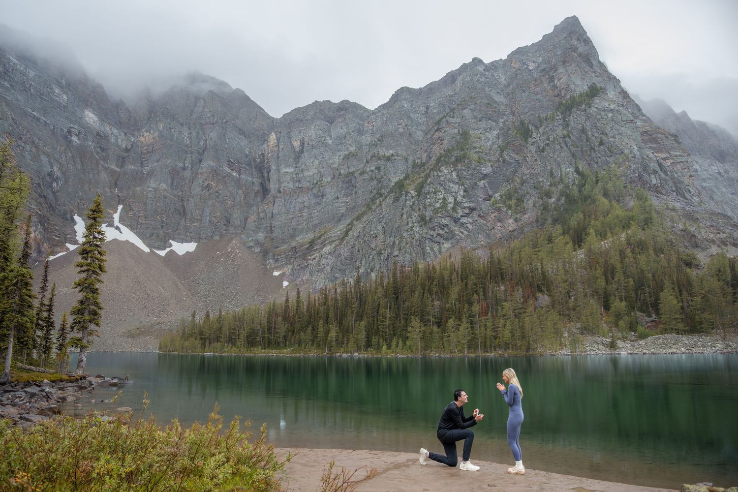 Man proposing to woman by a serene mountain lake with dramatic rocky peaks and evergreen trees in the background.