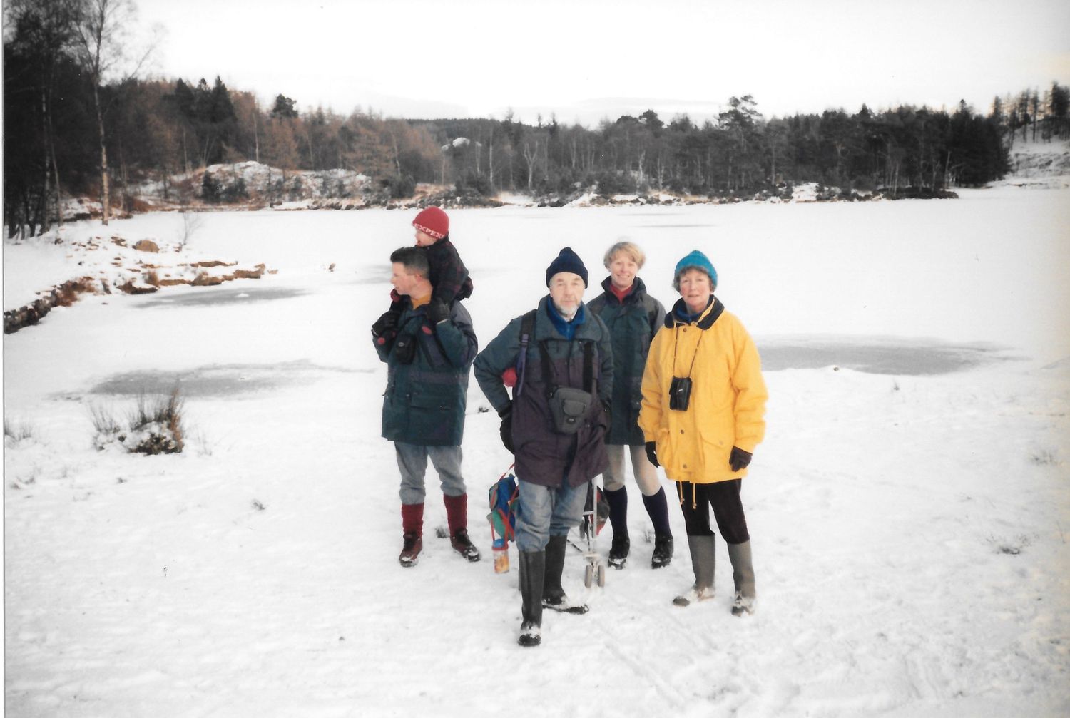 Group of five bundled up visitors posing outdoors in snowy winter scene near icy lake with forest backdrop.
