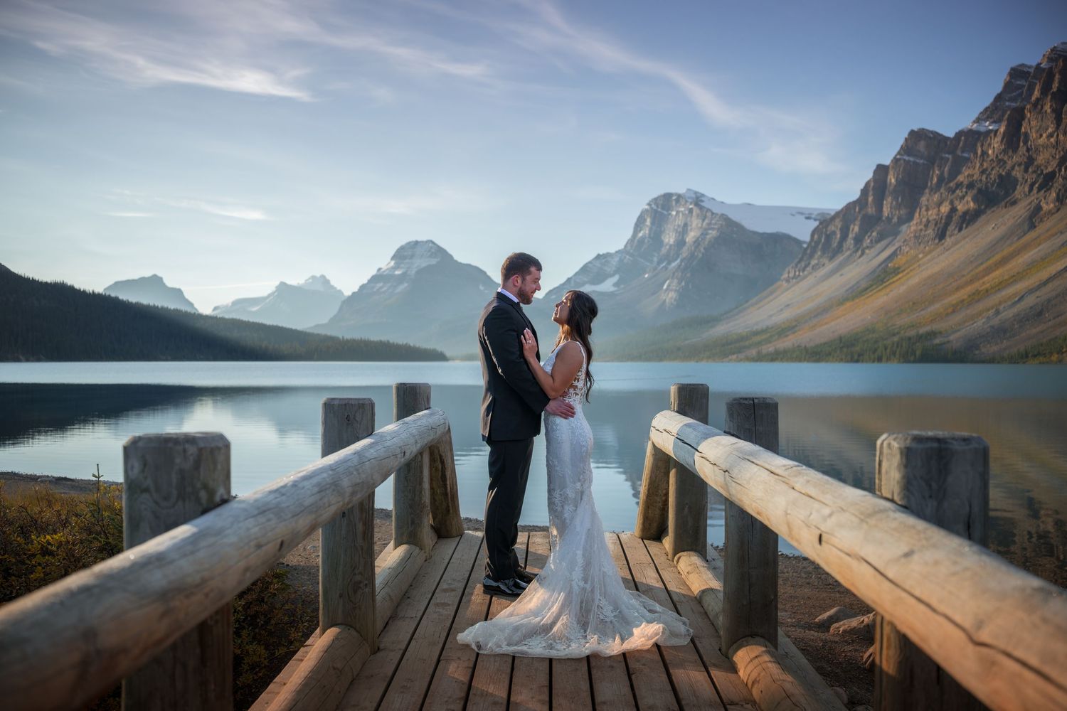 Bride and groom share a romantic moment on a wooden deck overlooking a serene mountain lake at sunset.