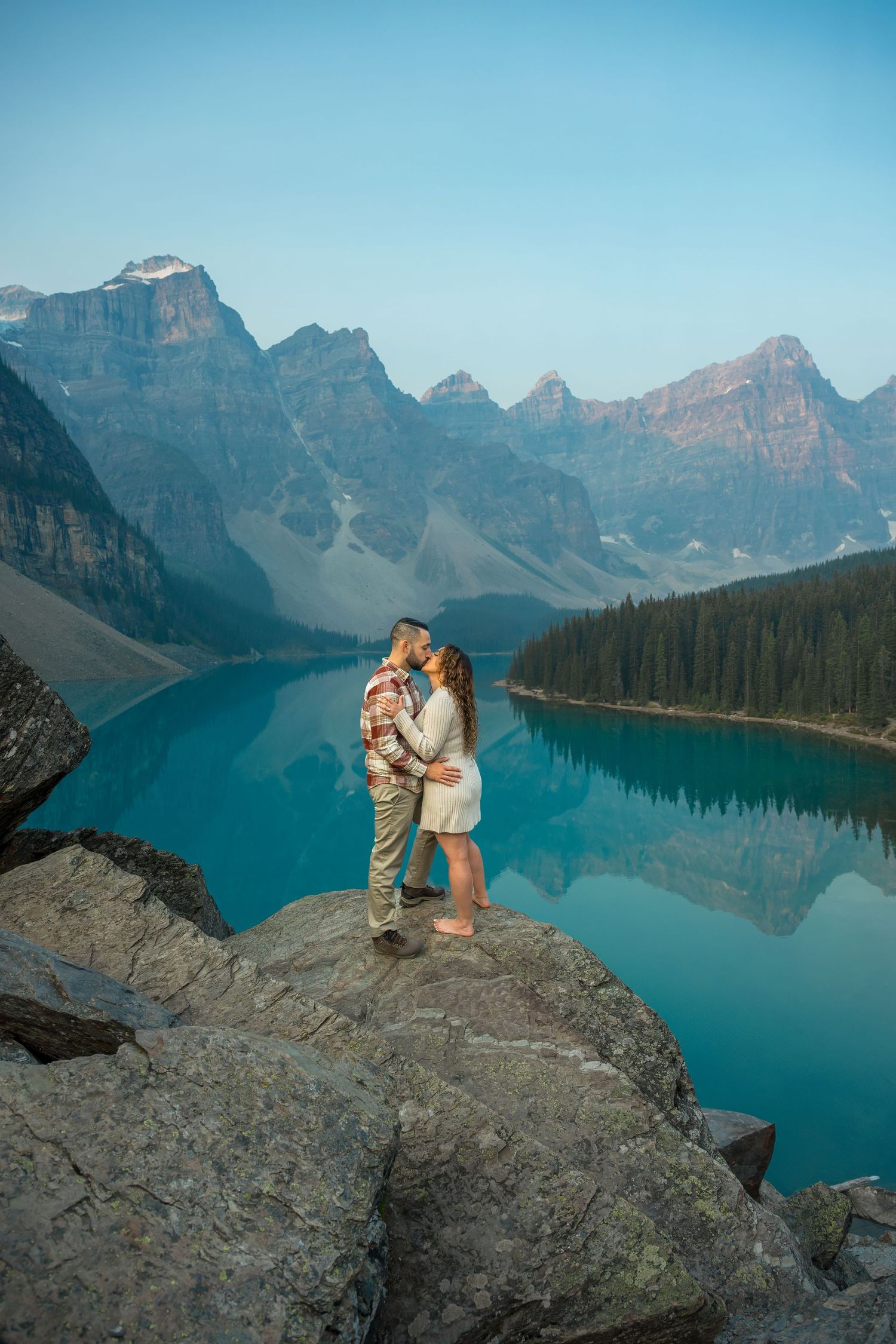 Romantic couple embracing on rocks above vivid turquoise Moraine Lake surrounded by Canadian Rockies.