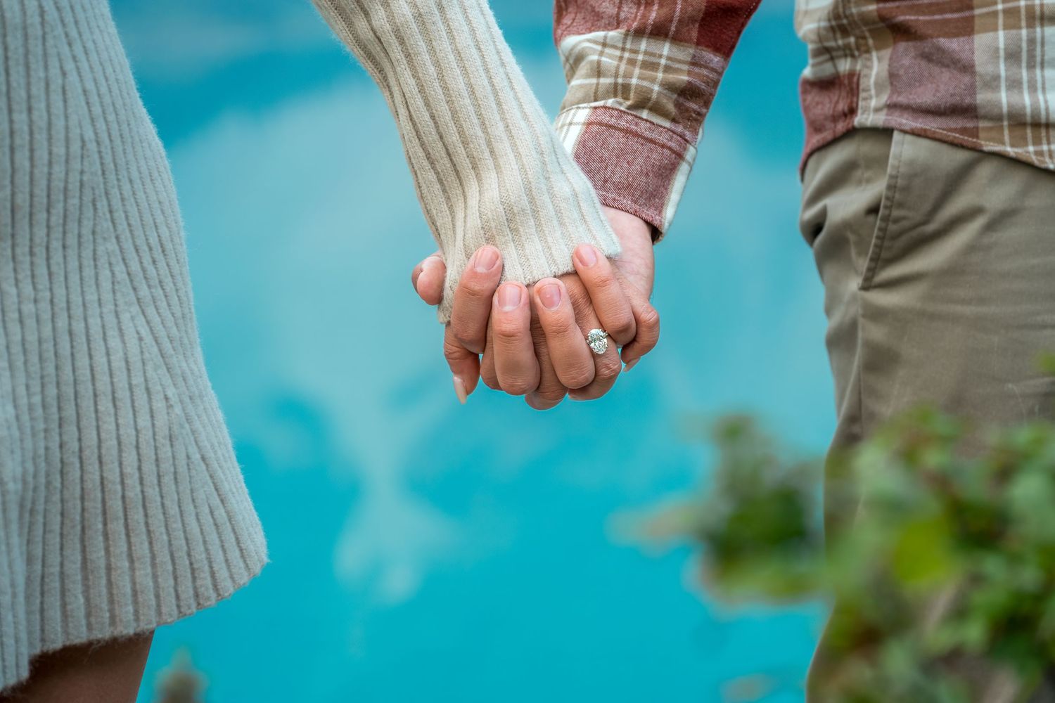 Close-up of couple holding hands with diamond engagement ring, turquoise Moraine Lake behind them.
