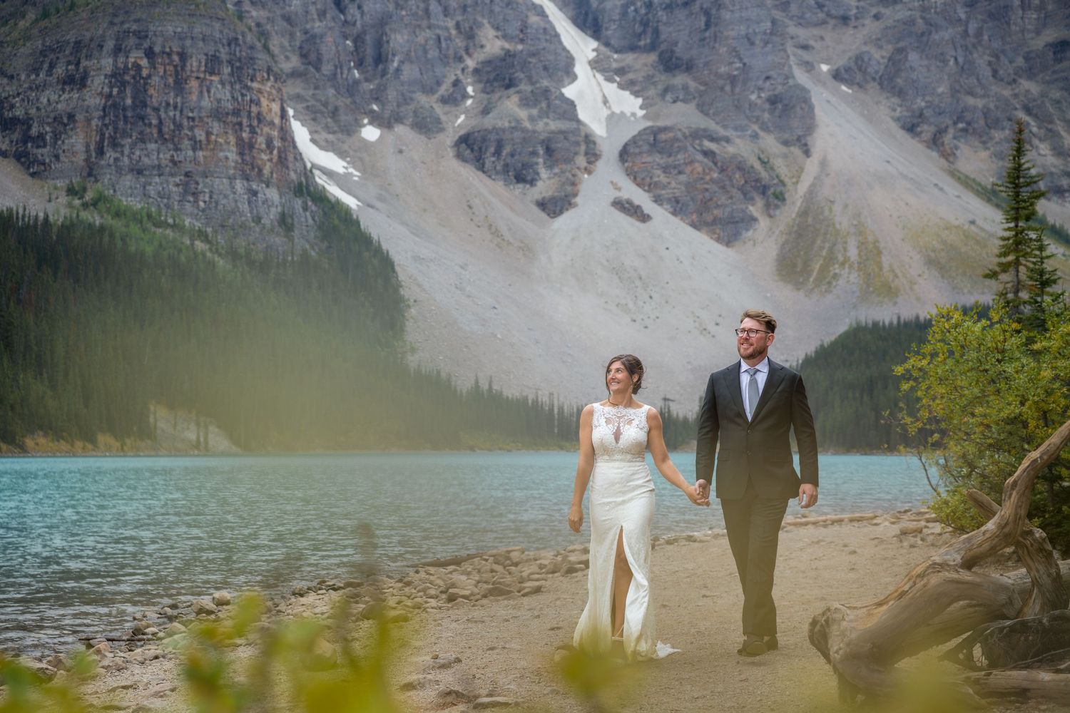 Bride and groom walk hand-in-hand on a rocky shore at Moraine Lake with turquoise water and mountains behind.