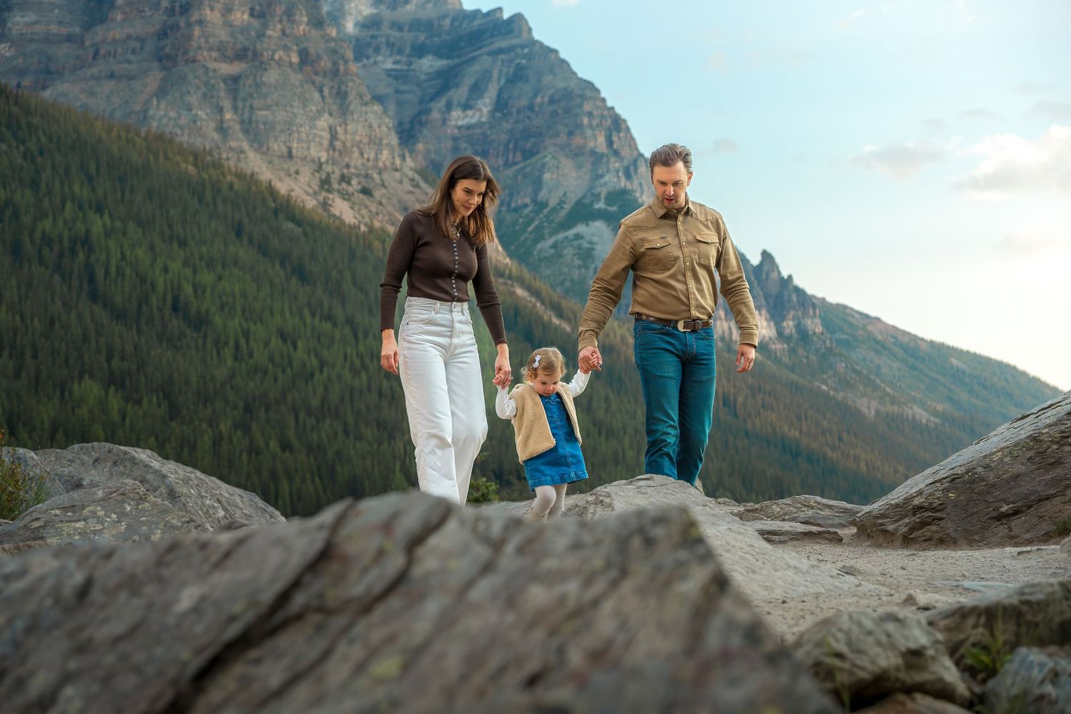 Family with young child walking on rocky terrain with mountain forest backdrop in Banff National Park.