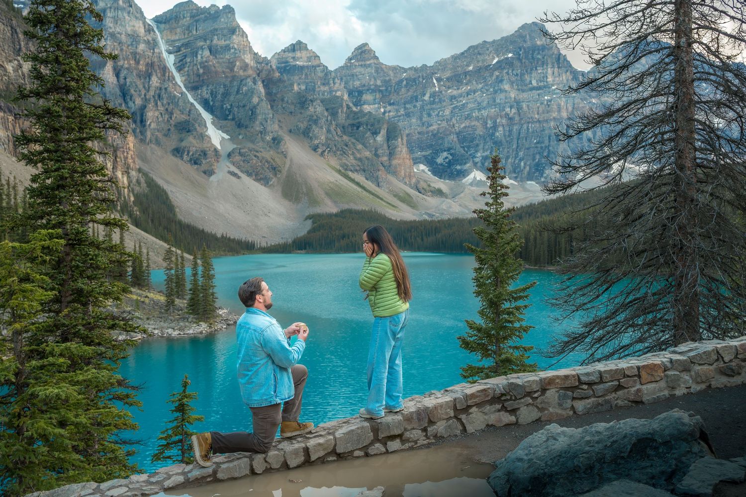 Man proposing on one knee to woman in green at scenic Moraine Lake with Rocky Mountain views.