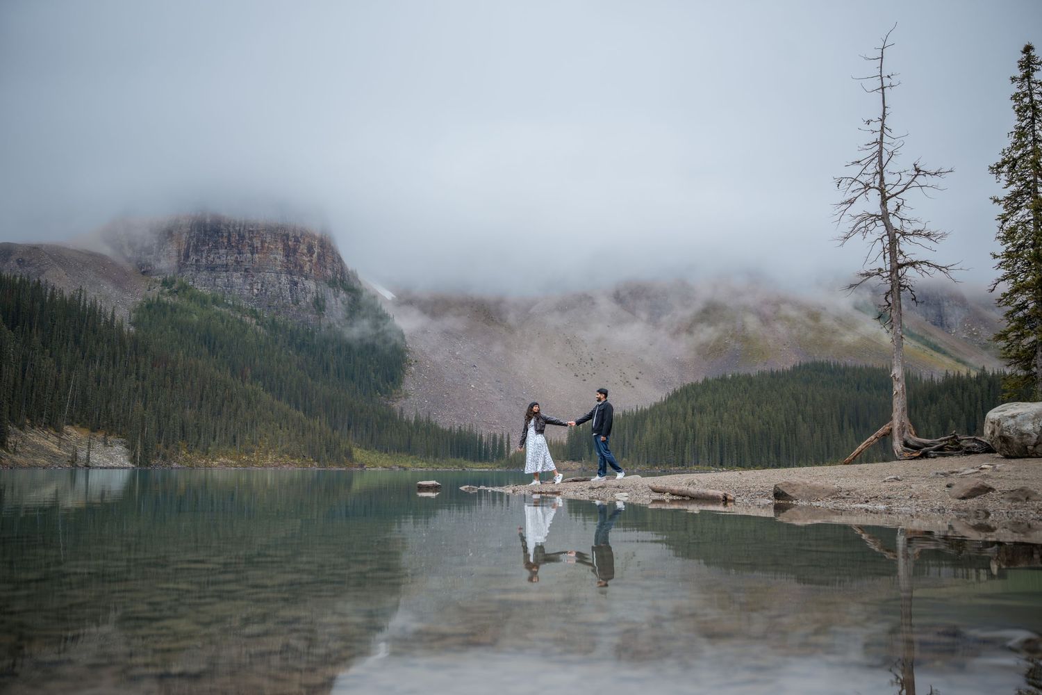 Couple holds hands by a misty mountain lake with forest reflections in Banff National Park during their elopement.