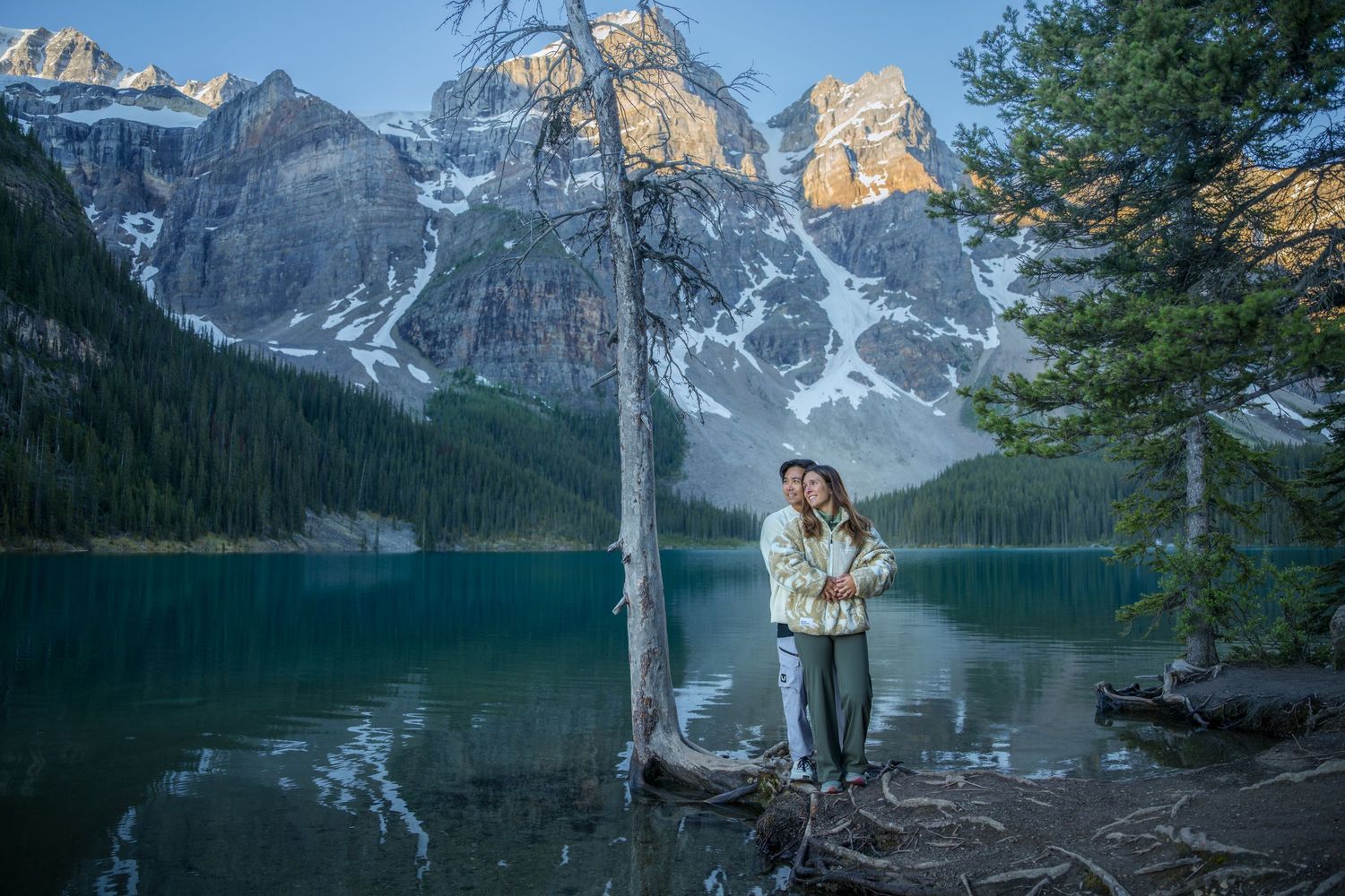 Couple embracing by turquoise Moraine Lake with snow-capped Rocky Mountains reflected in water, Banff National Park, Canada.