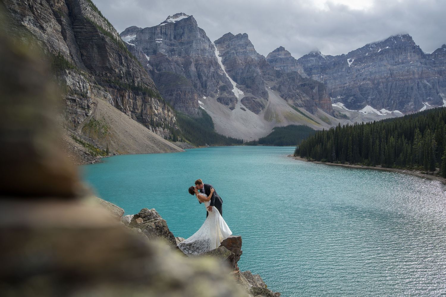 Couple sharing a romantic kiss on rocky cliffs at Moraine Lake, Banff, with stunning turquoise water and mountains.