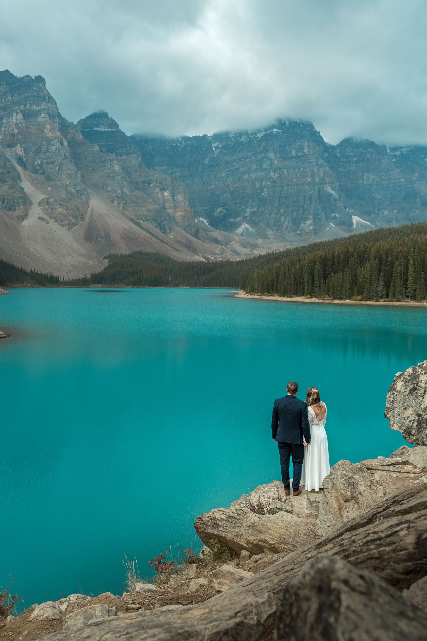Bride and groom standing on rocky ledge overlooking vivid turquoise Moraine Lake and Rocky Mountains.