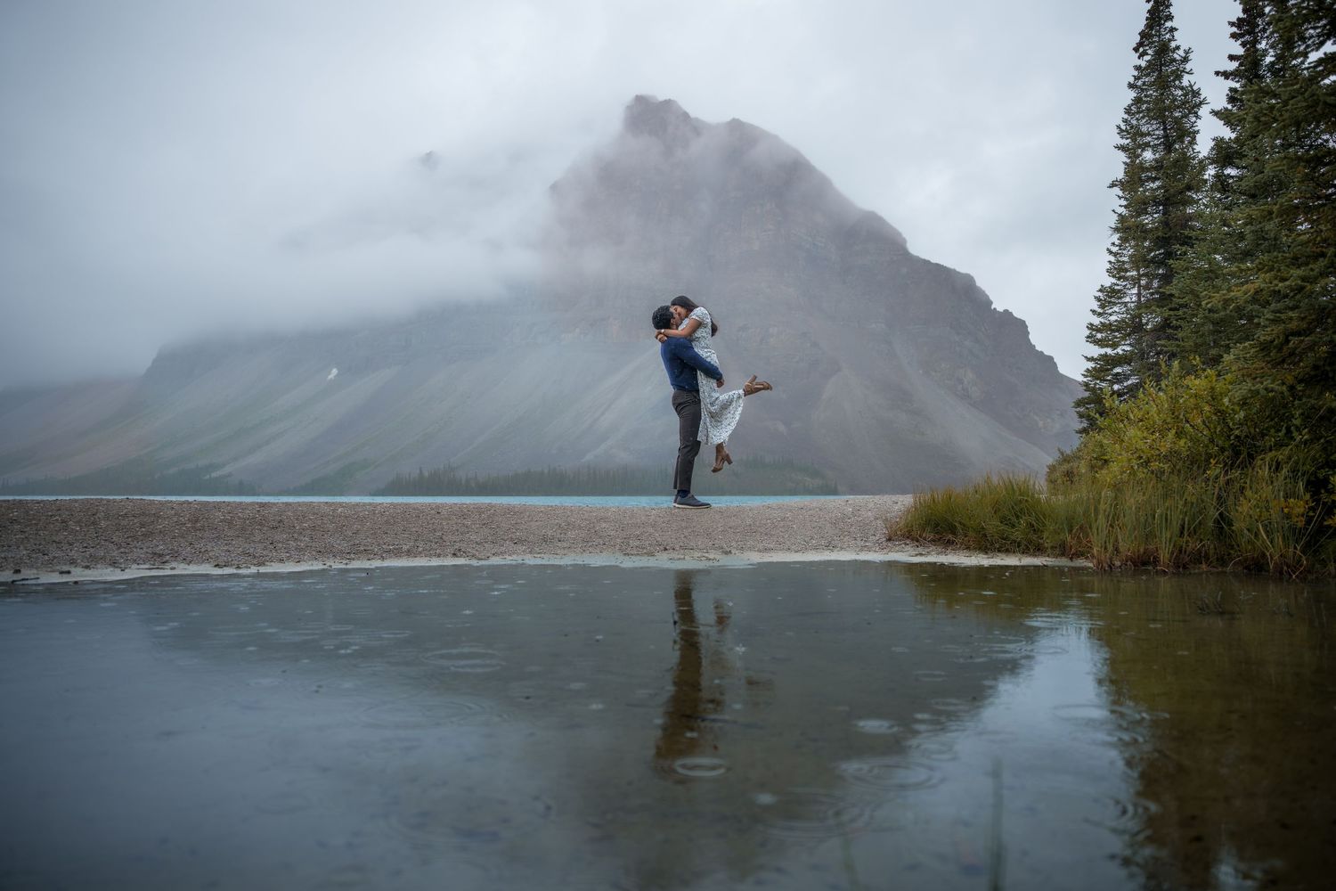 Couple embracing by a misty mountain lake in Banff, Canada, with stunning rocky peaks and evergreen trees reflected in calm water.