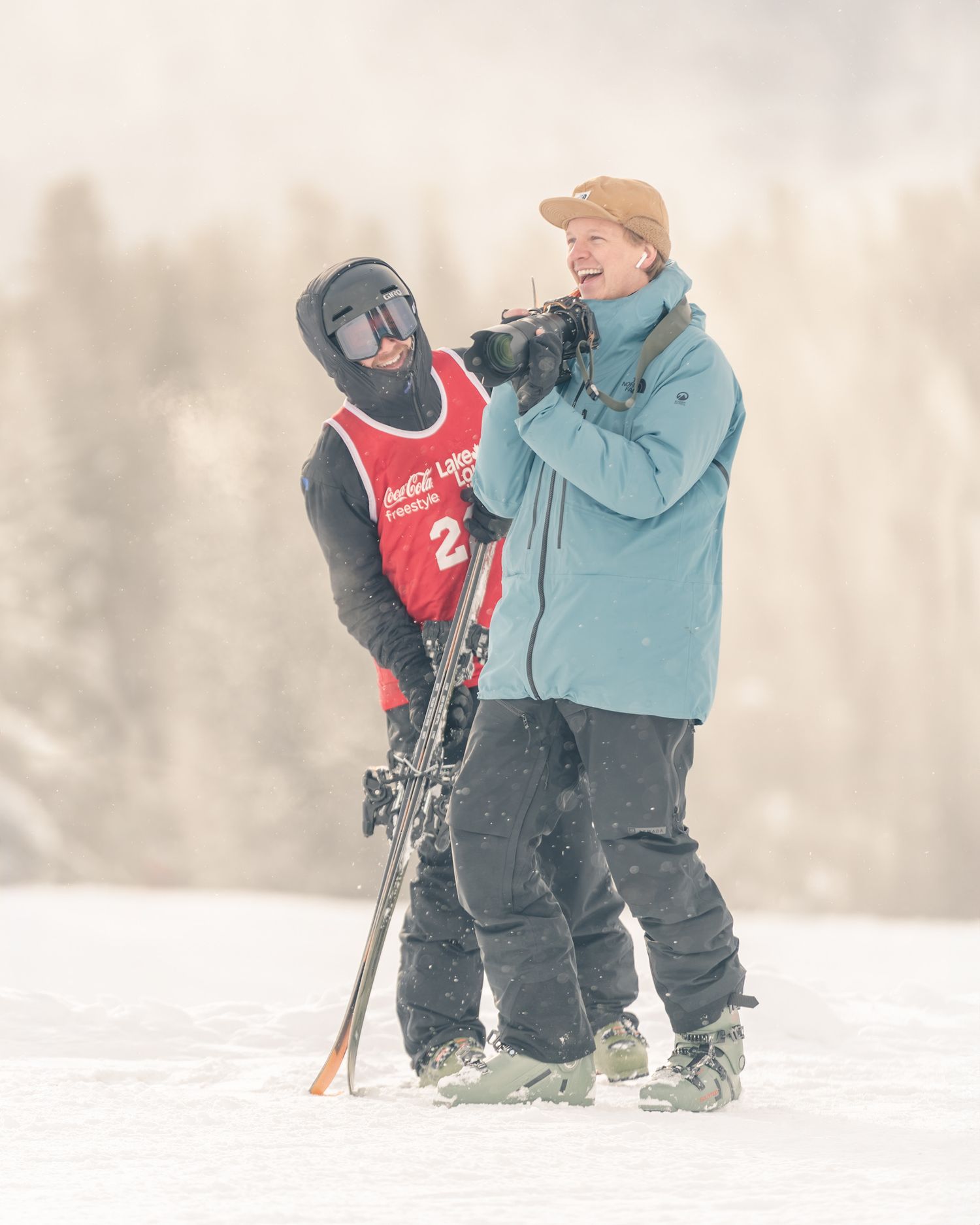 Two skiers standing in snowfall on ski slope, one filming with camera equipment.