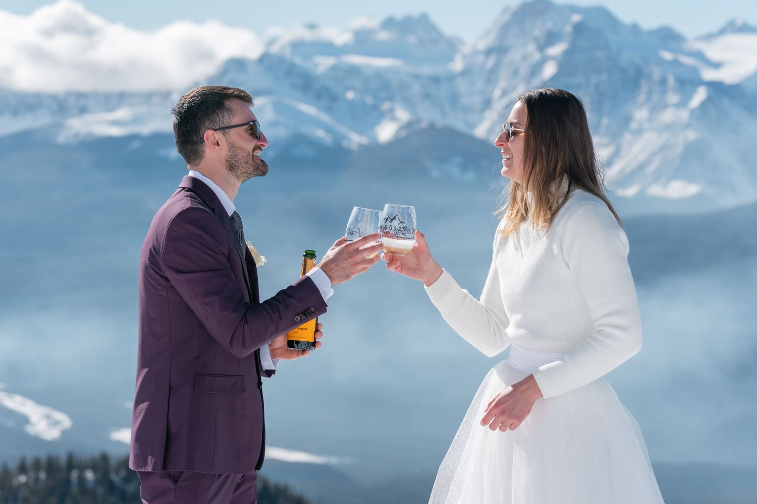 Couple in wedding attire toasting champagne glasses on snowy mountain summit with stunning alpine peaks backdrop.