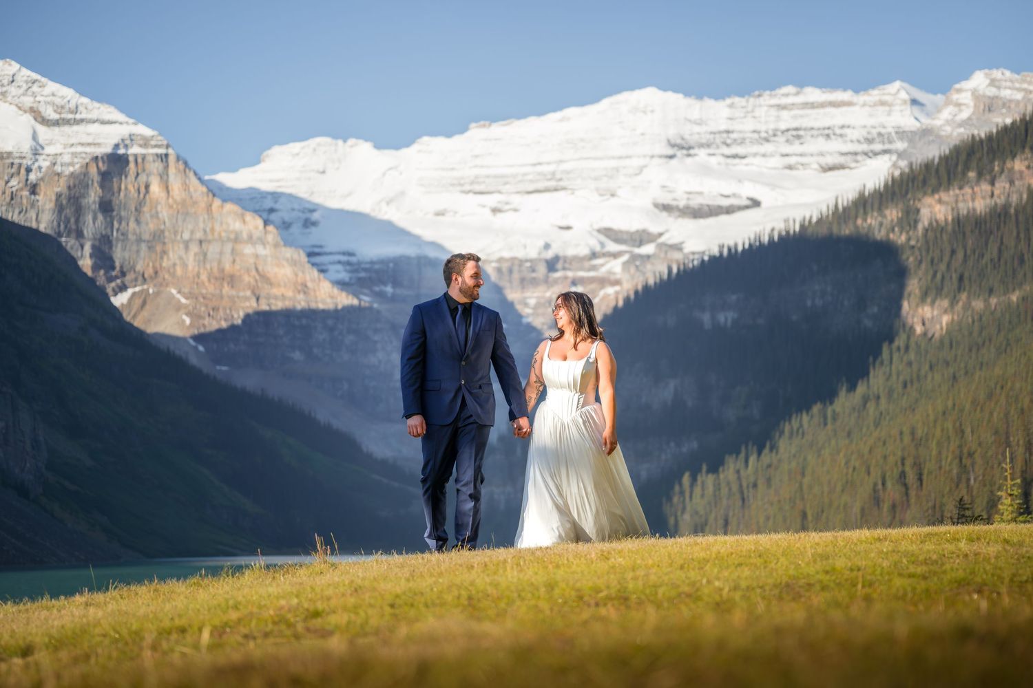 Bride and groom holding hands walking in a mountain meadow with snow-capped peaks and lake in background.