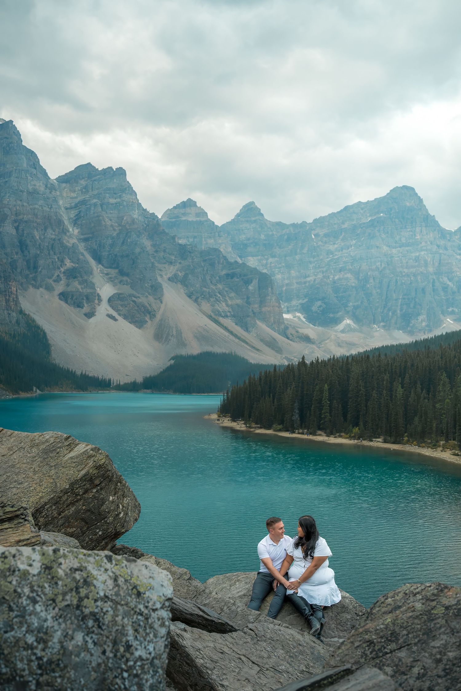 Couple sitting together on rocks at Moraine Lake with turquoise water and mountain scenery behind them.