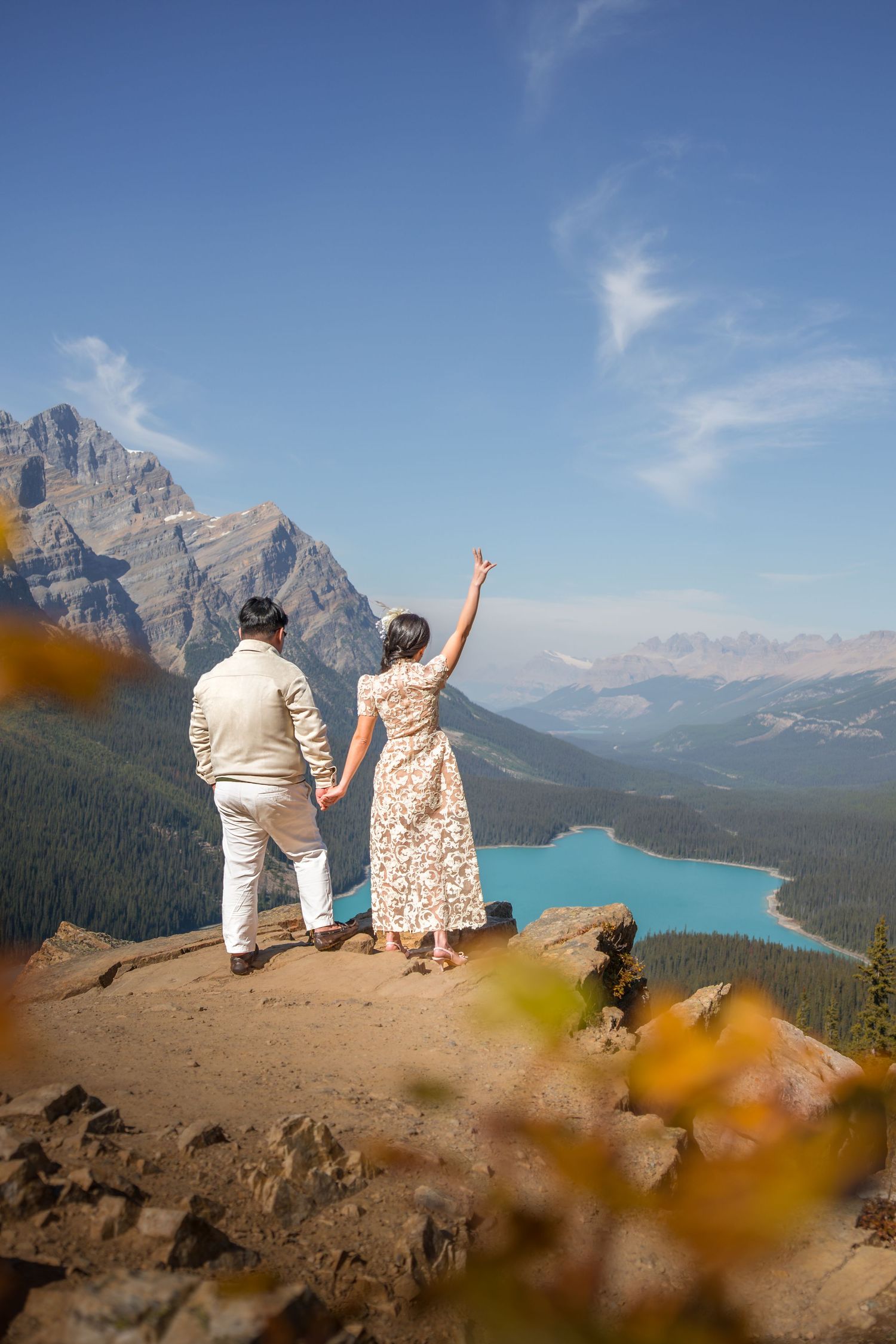 Couple holding hands on rocky overlook above turquoise Peyto Lake, Canadian Rockies, woman raising arm in celebration.