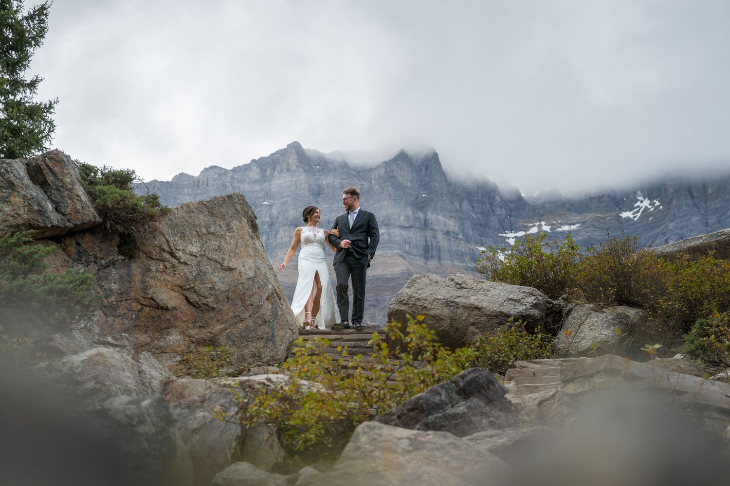 Bride and groom stand on rocky mountain steps with misty peaks behind them during an outdoor elopement ceremony.