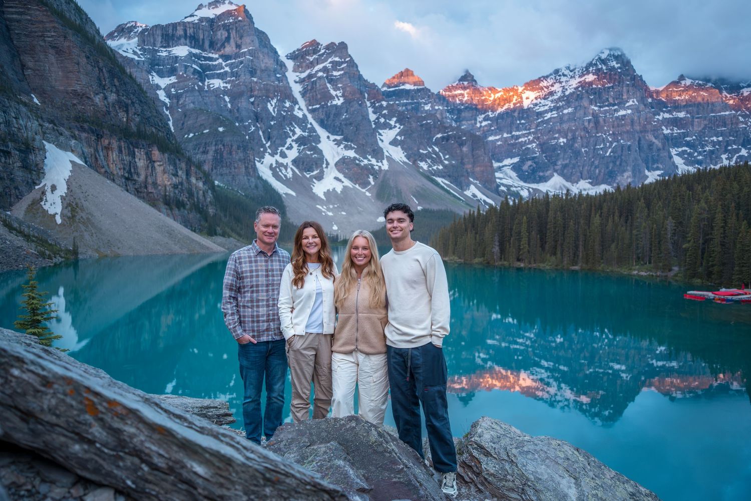 Family of four smiling at Moraine Lake, Banff, with turquoise water and snow-capped Rocky Mountains at sunset.