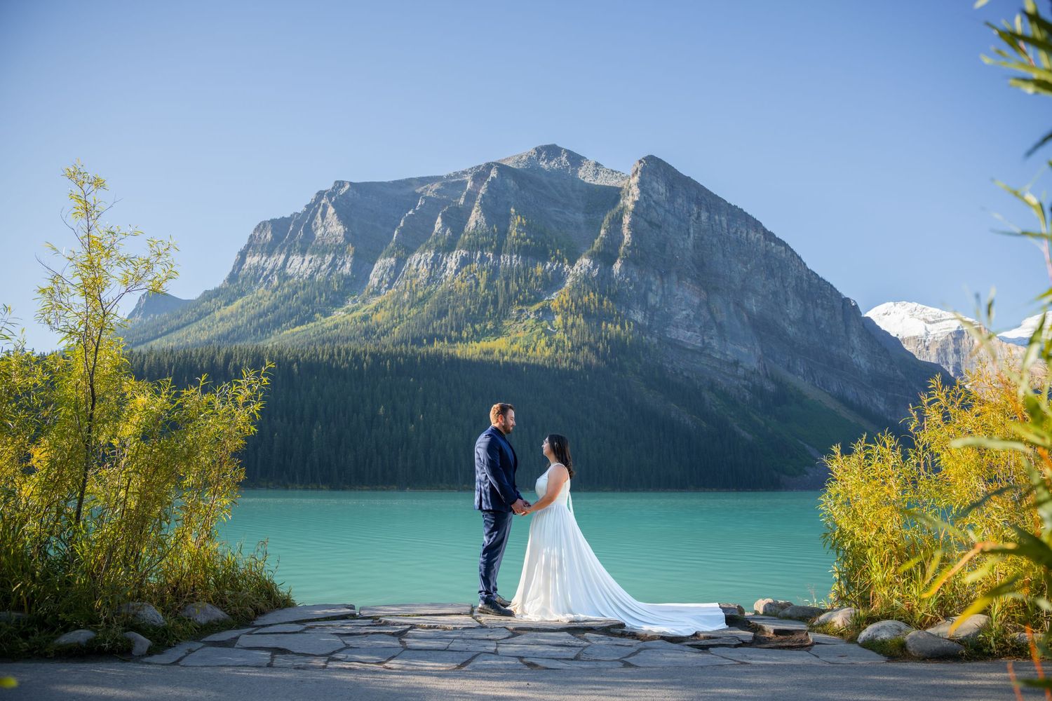Bride and groom stand before turquoise Lake Louise with majestic Rocky Mountain peaks in Banff, Alberta, Canada.