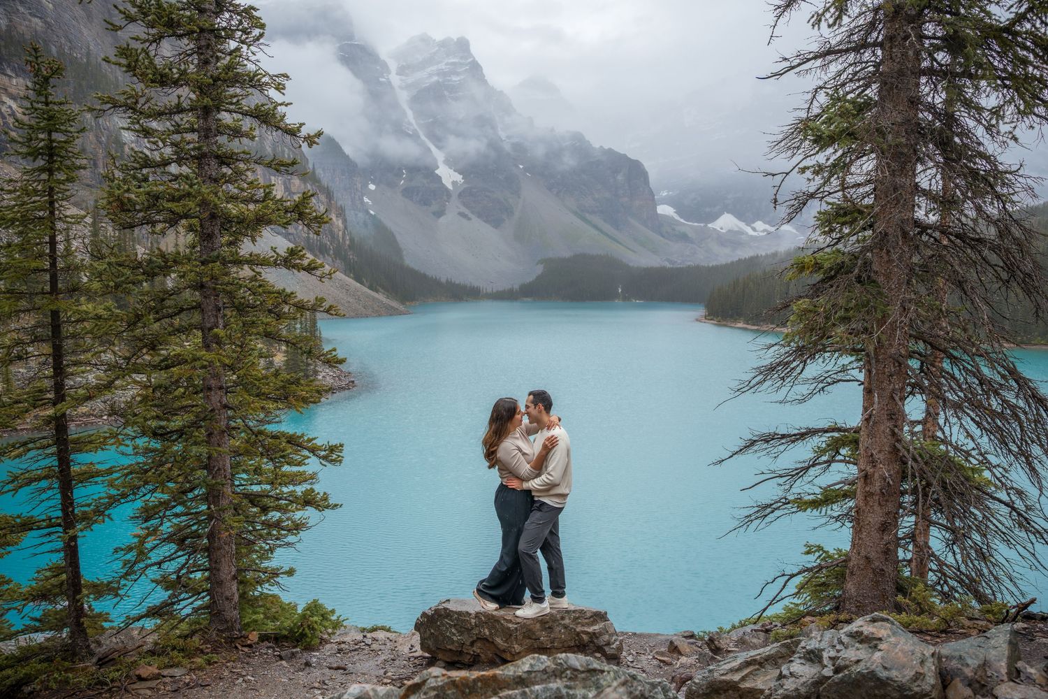 Couple embracing on rocks at turquoise Moraine Lake, Banff, surrounded by evergreens and snow-capped mountains.