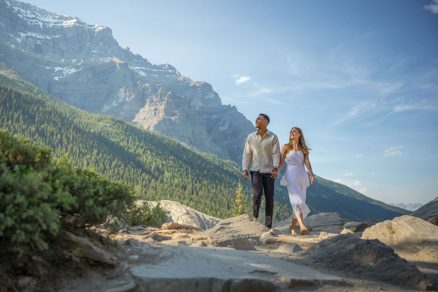 Couple holding hands walking on rocky mountain trail with stunning peaks and evergreen forest backdrop on sunny day.