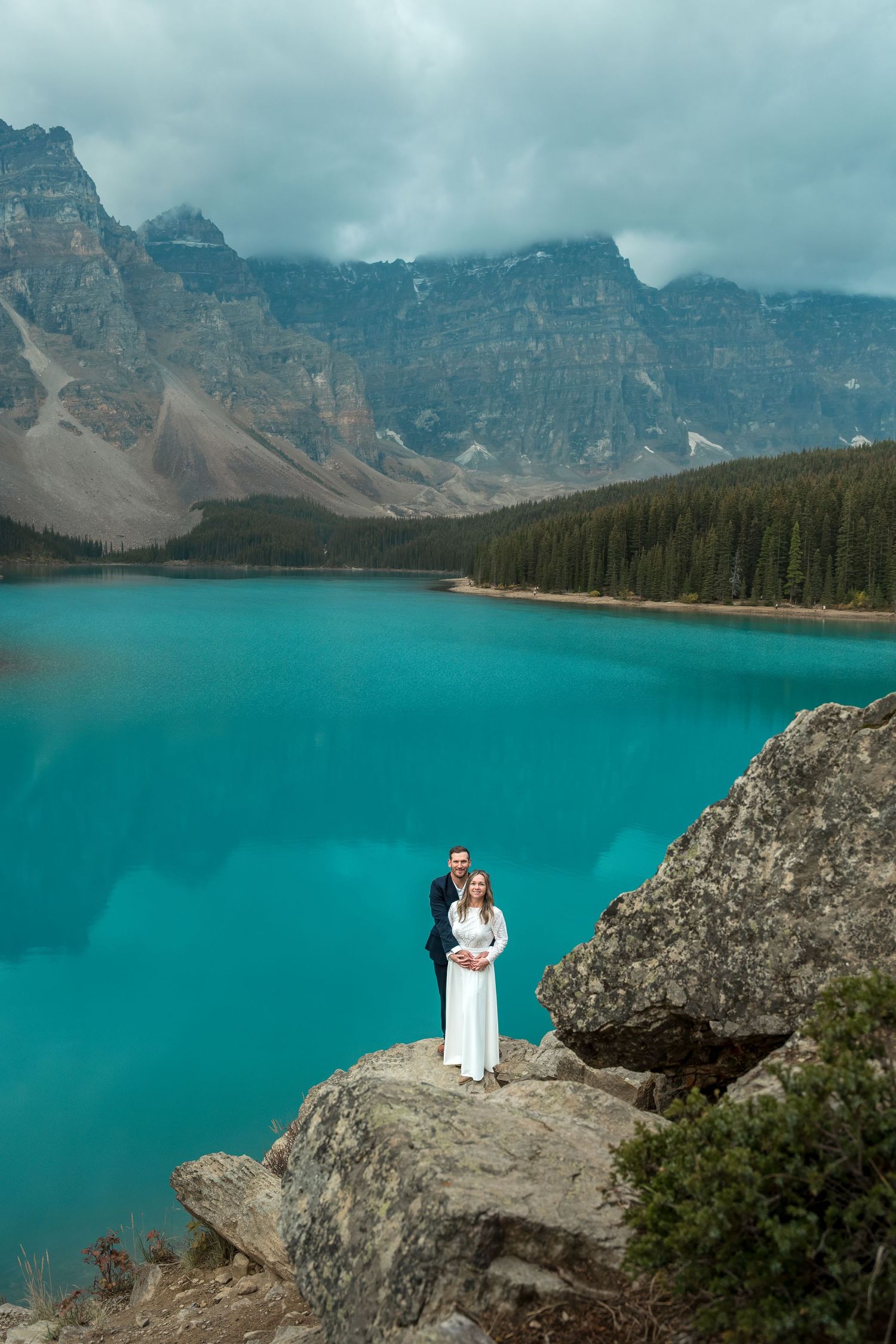 Couple in wedding attire standing on rocky ledge overlooking turquoise Moraine Lake with mountains in Banff, Canada.