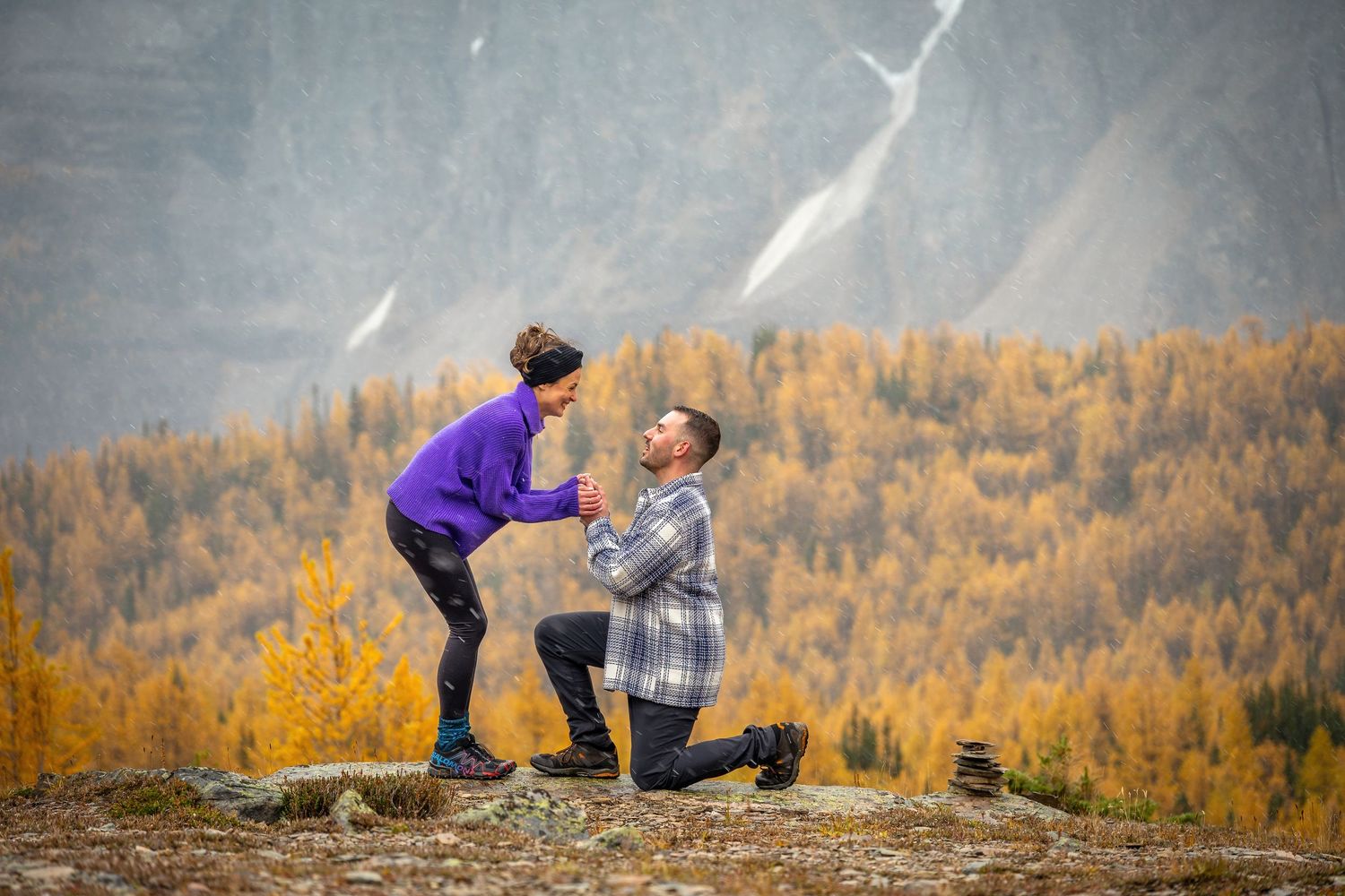 Man kneeling on mountain overlook proposing to woman with colorful autumn forest backdrop.