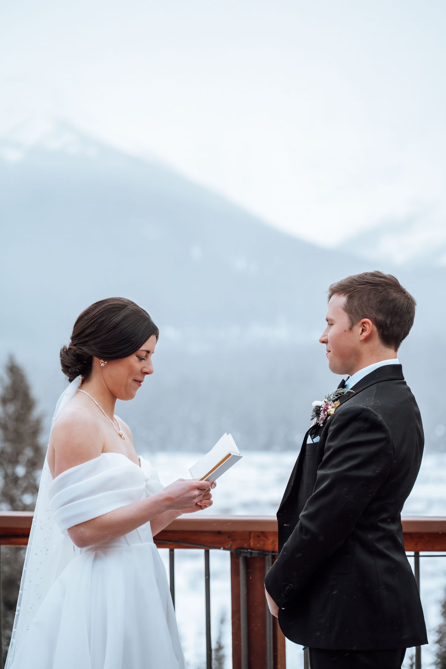 Bride reads wedding vows to groom on snowy mountain balcony, wearing white dress and fur wrap during winter ceremony.