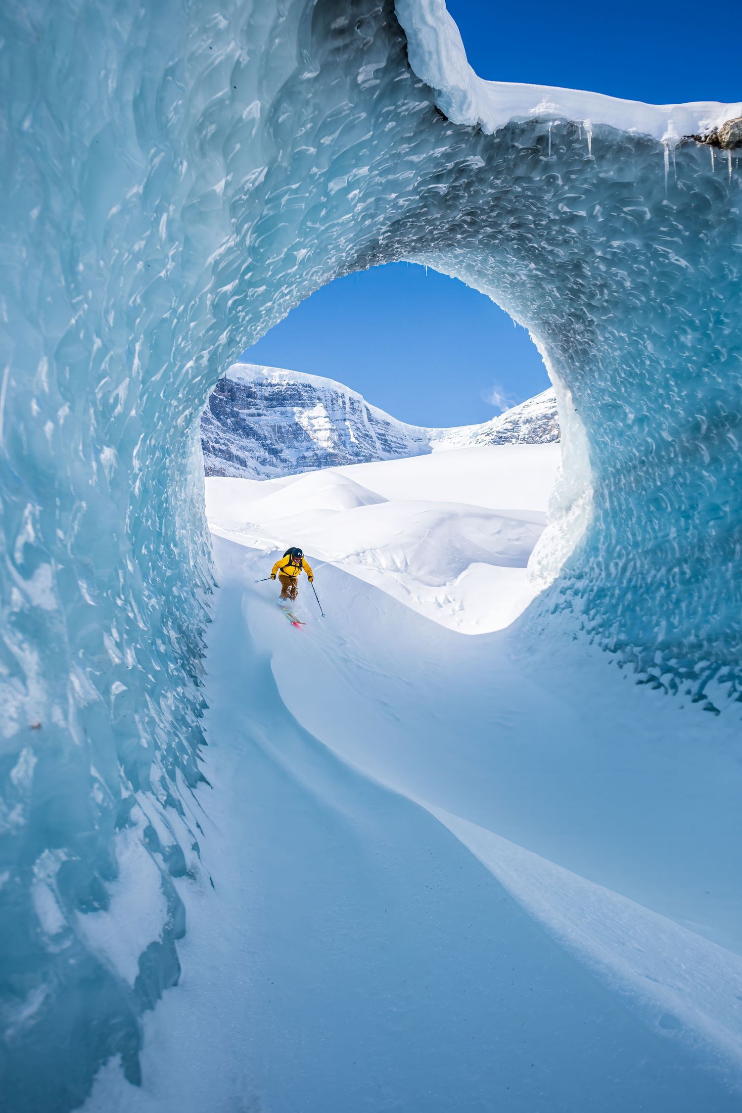 Skier in yellow jacket skiing through dramatic blue ice cave tunnel on snowy glacier.