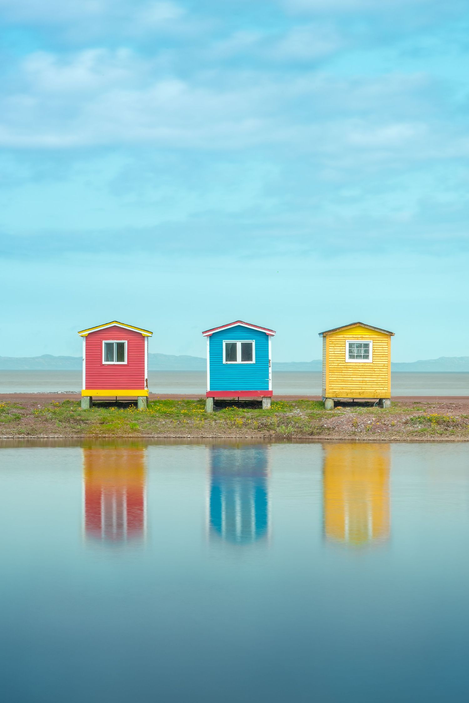 Three colorful red, blue, and yellow wooden huts reflecting in calm water under blue sky.