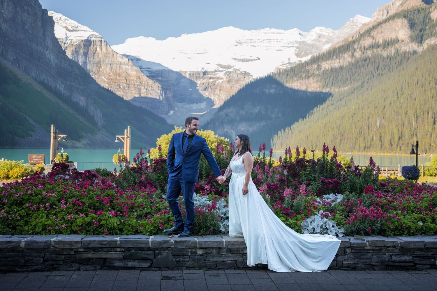 Bride and groom hold hands at Lake Louise with stunning mountain backdrop and colorful flowers at Fairmont Chateau.