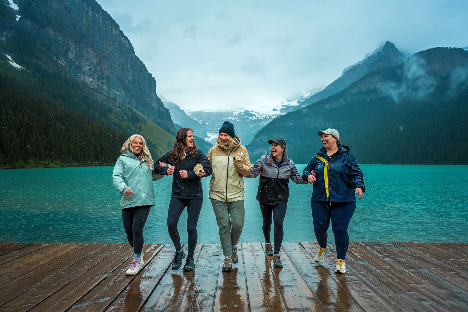 Five women laughing and walking arm-in-arm on a dock at turquoise Lake Louise on a rainy day.