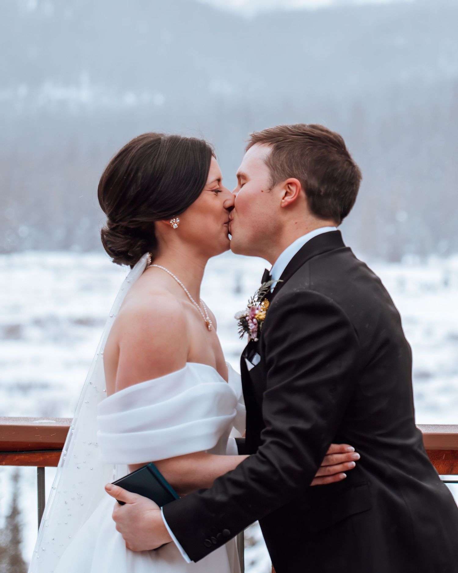 Bride and groom share a kiss outdoors in a snowy winter mountain setting, bride in white dress with wrap.