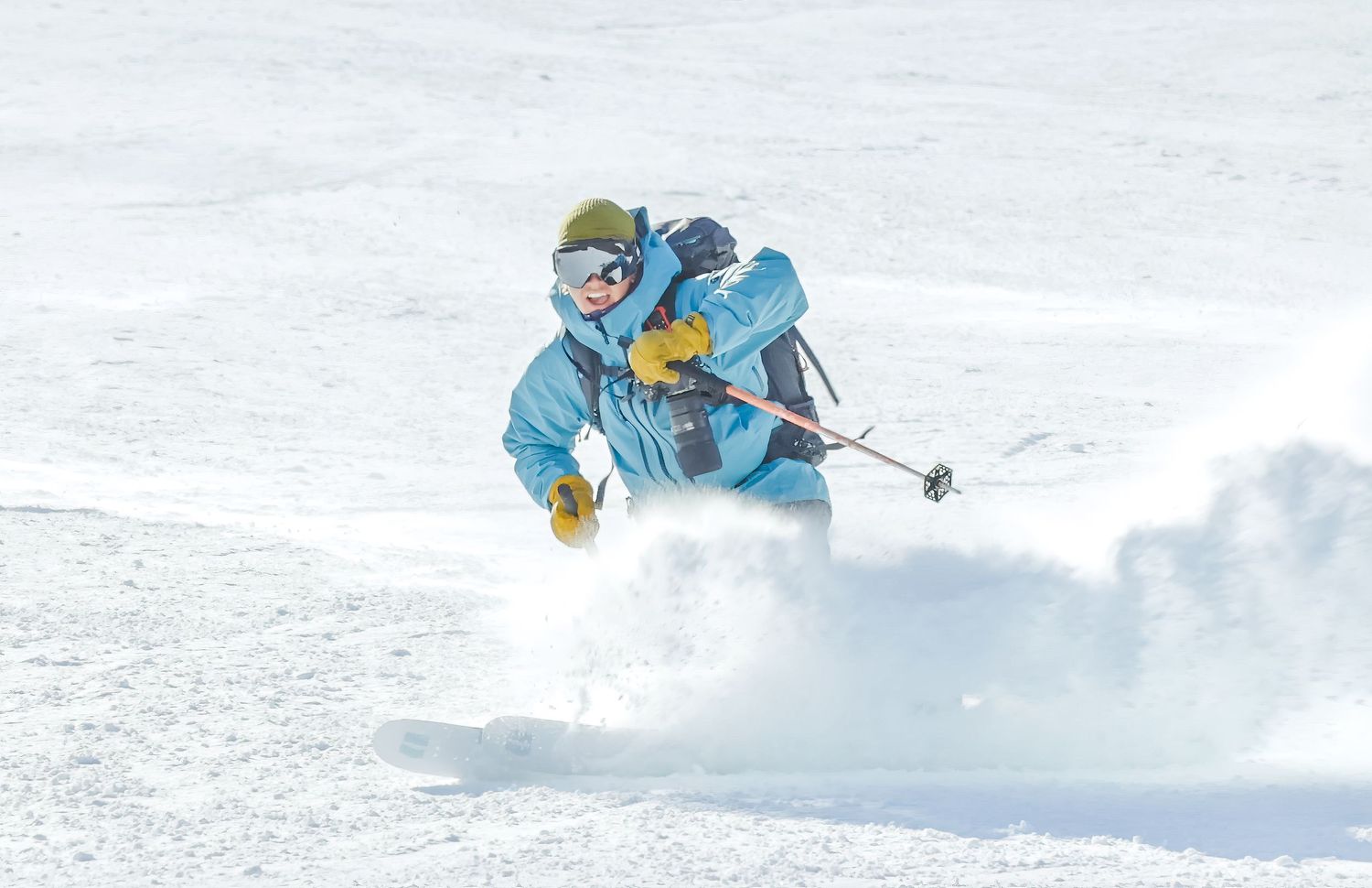 Skier in blue jacket carving through deep powder snow on a bright sunny mountain slope.