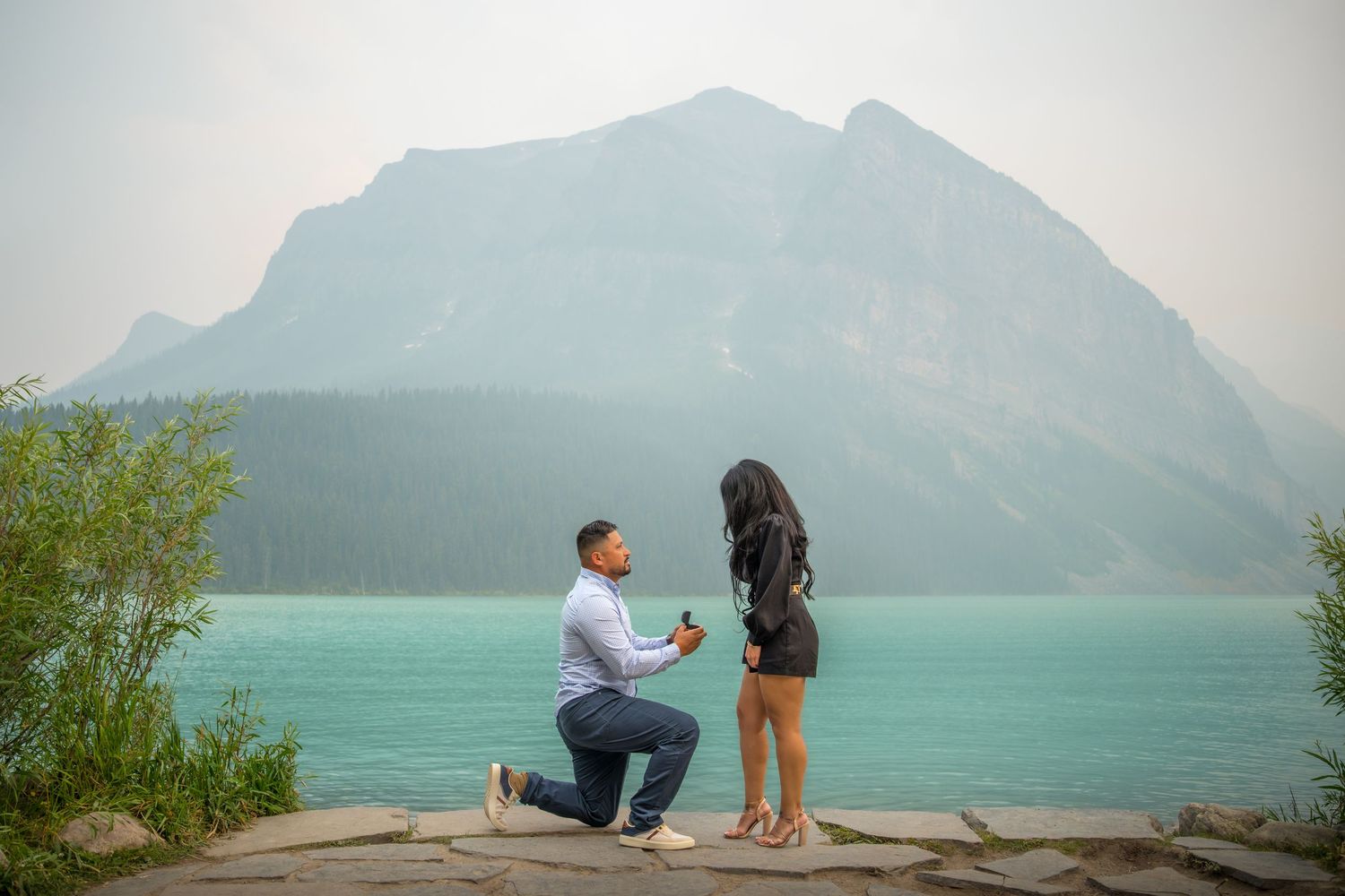 Man proposes on one knee by turquoise lake with mountain backdrop in Banff, Canada.