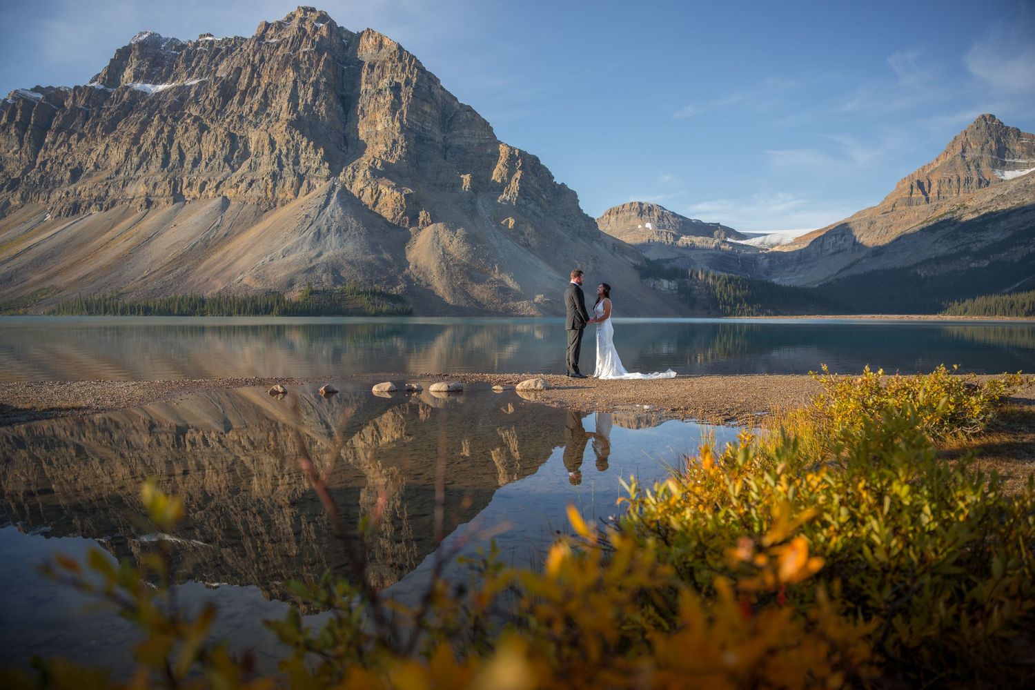 Bride and groom stand by a reflective mountain lake with stunning rocky peaks in Banff National Park during fall.