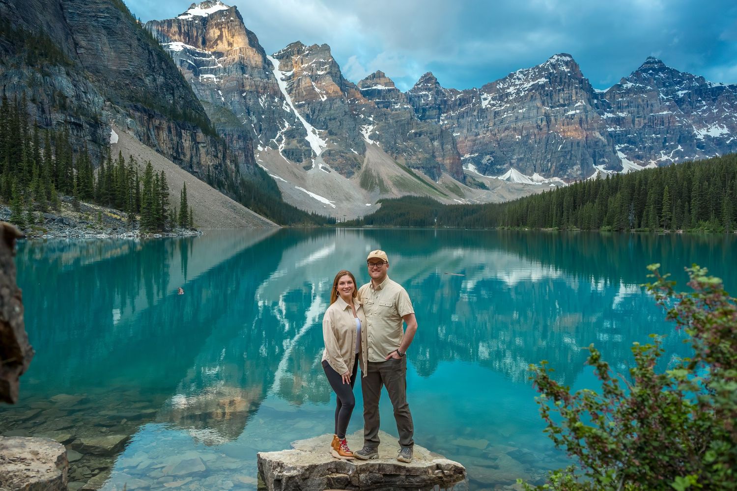 Couple standing on rocks at turquoise Moraine Lake with snow-capped Rocky Mountains reflected in the water.