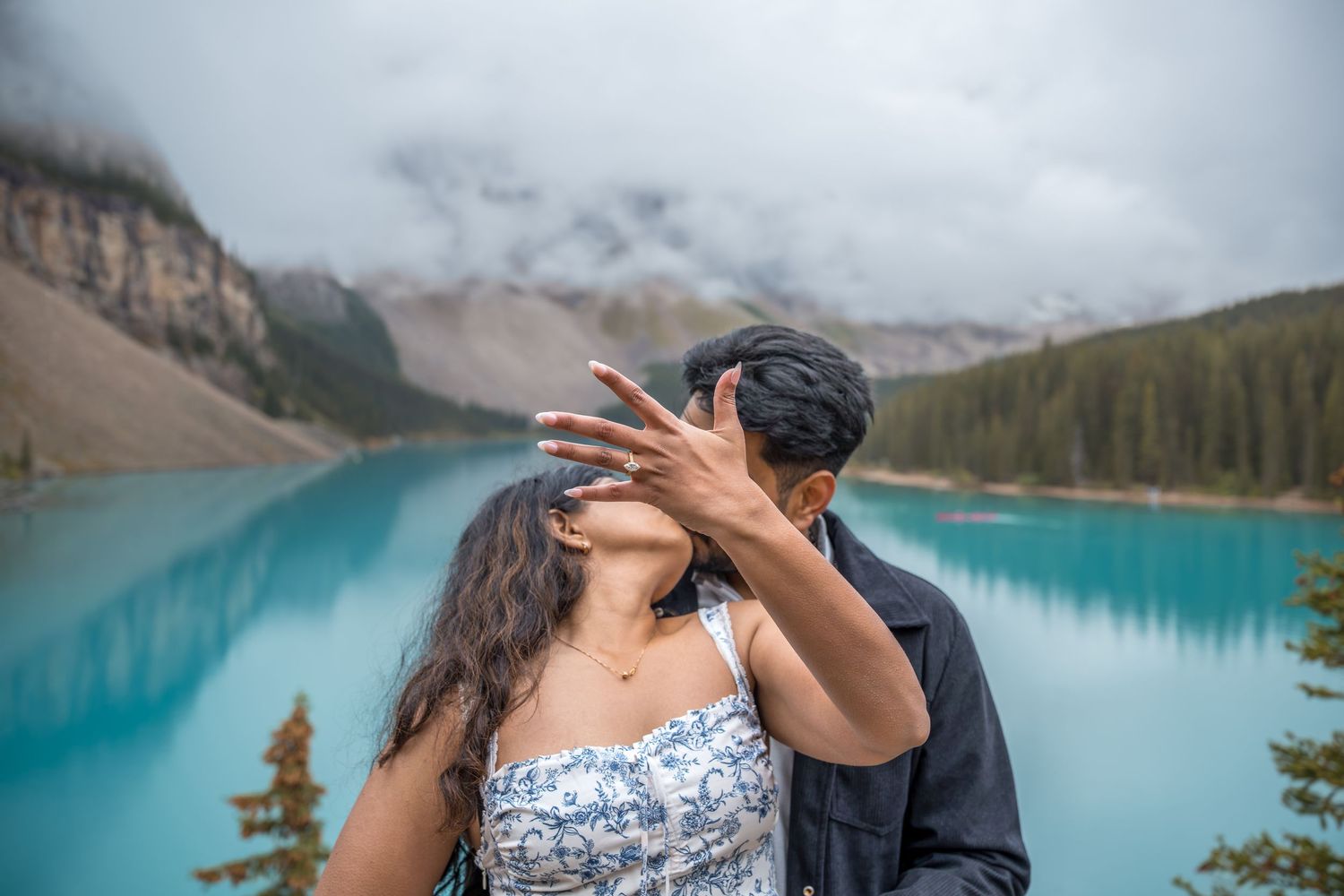 Couple sharing a kiss at Moraine Lake, Canada, with turquoise waters and misty mountains in the background.