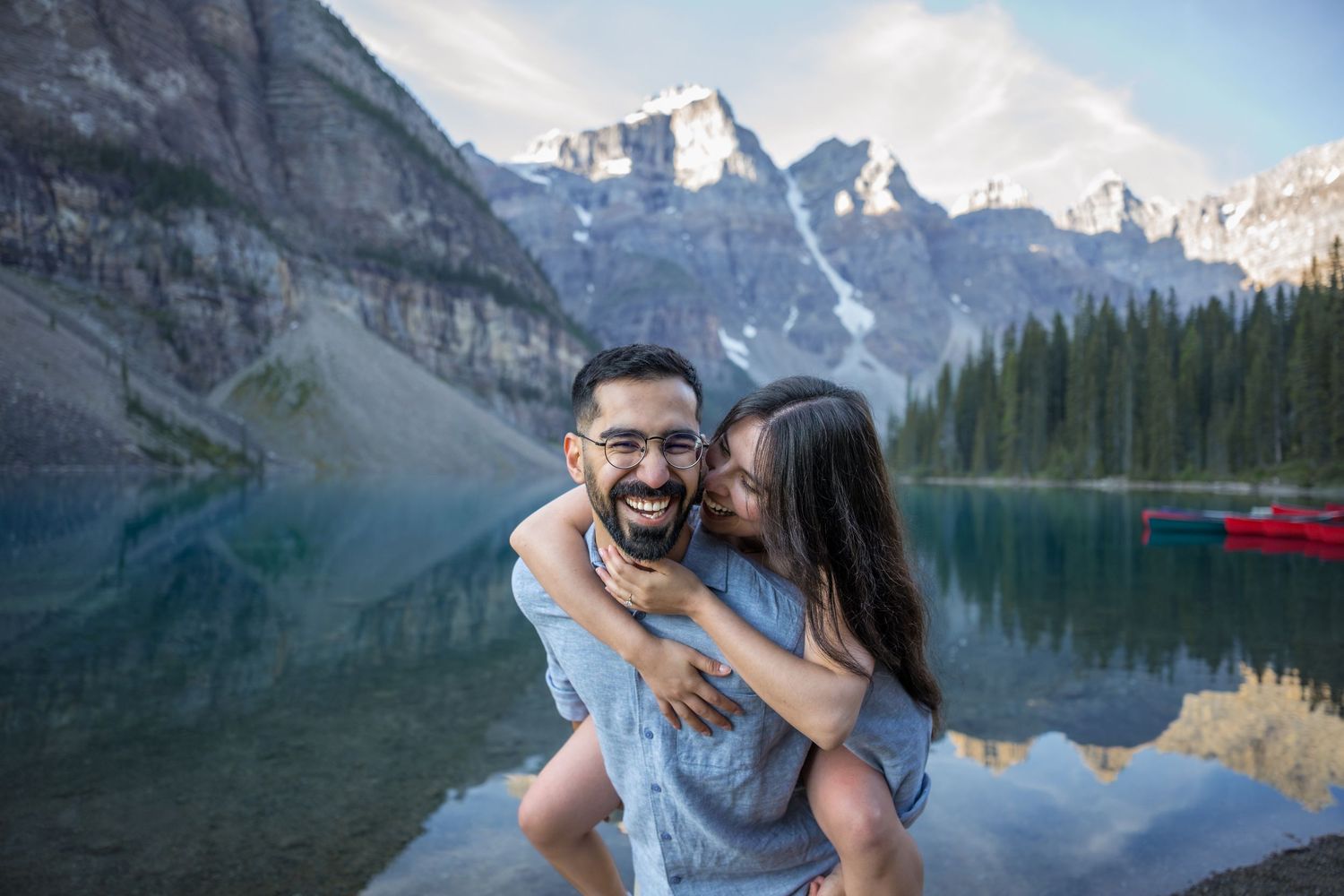 Couple laughing at Moraine Lake, Banff, woman kissing man on cheek, mountains and turquoise water behind them.