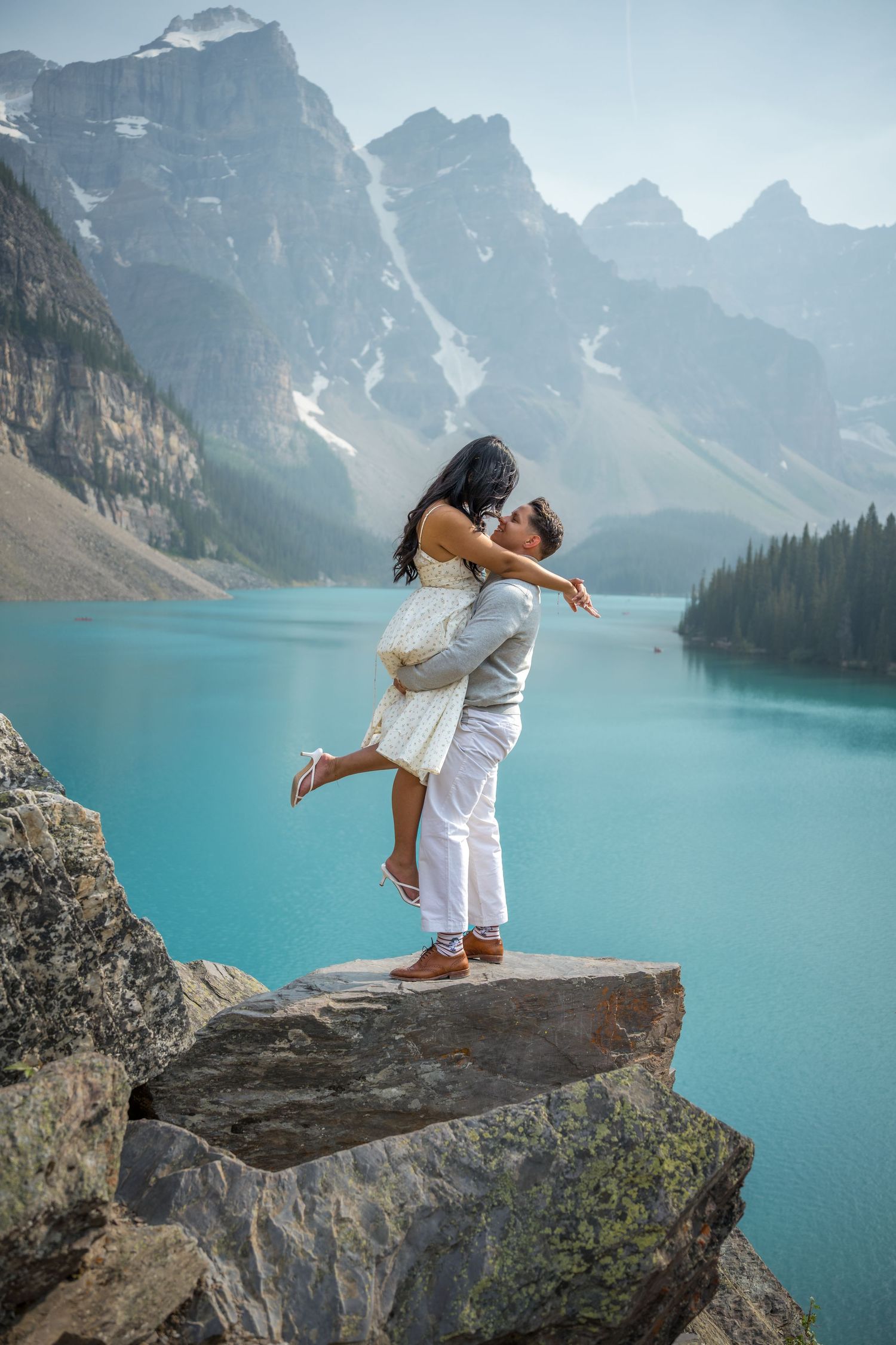 Couple embracing on rocky ledge at turquoise Moraine Lake with misty Canadian Rockies mountains in background.
