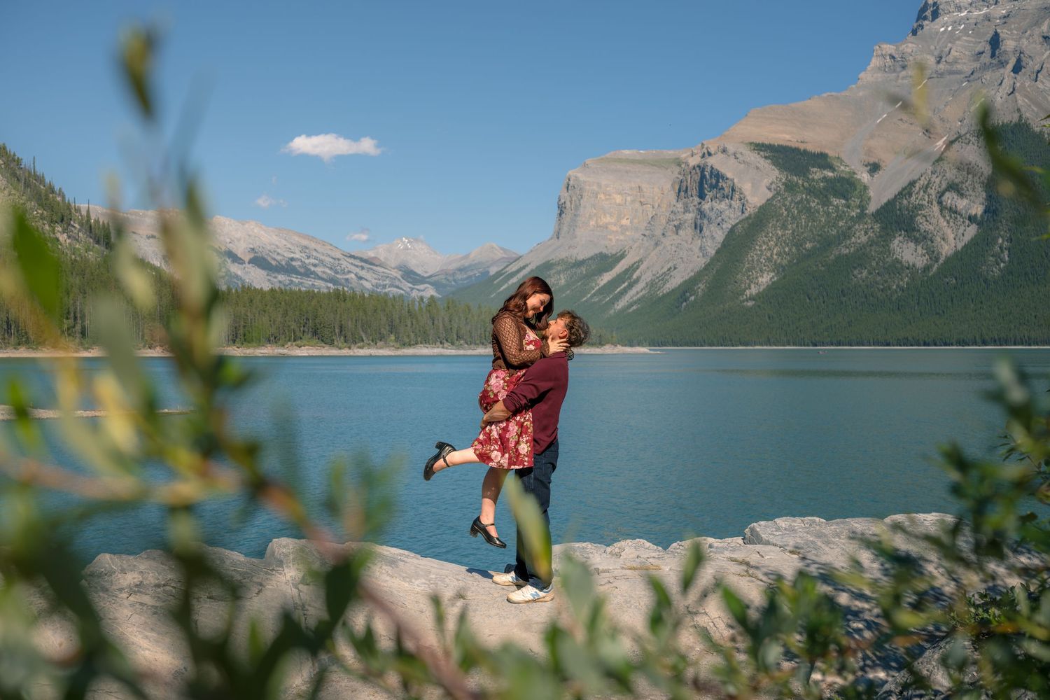 Couple embracing by turquoise Lake Minnewanka with Rocky Mountain peaks under blue sky in Banff National Park.