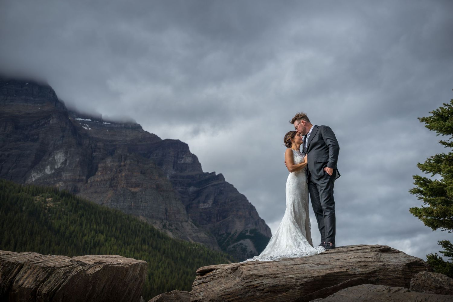 Bride and groom share a romantic kiss on rocky mountain overlook with dramatic cloudy peaks in Banff National Park.