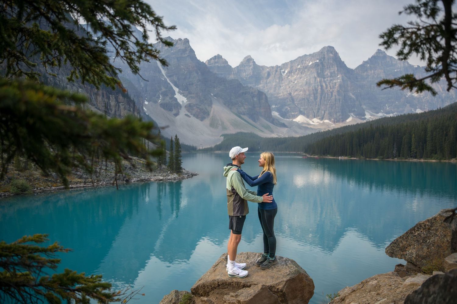 Couple standing on a rock embracing at turquoise Moraine Lake with Rocky Mountain peaks in Banff, Canada.