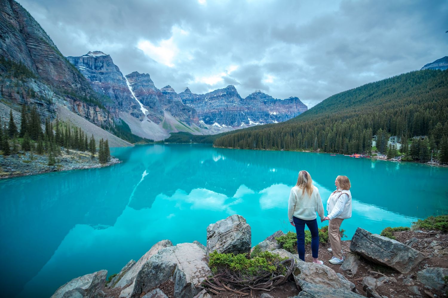 Two women stand on rocks overlooking turquoise Moraine Lake with dramatic Rocky Mountain peaks in Banff, Canada.