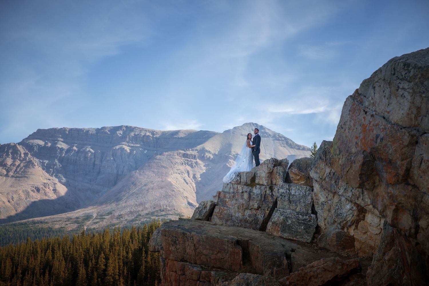 Couple standing on rocky mountain summit with stunning snow-capped peaks and blue sky backdrop during wedding elopement.