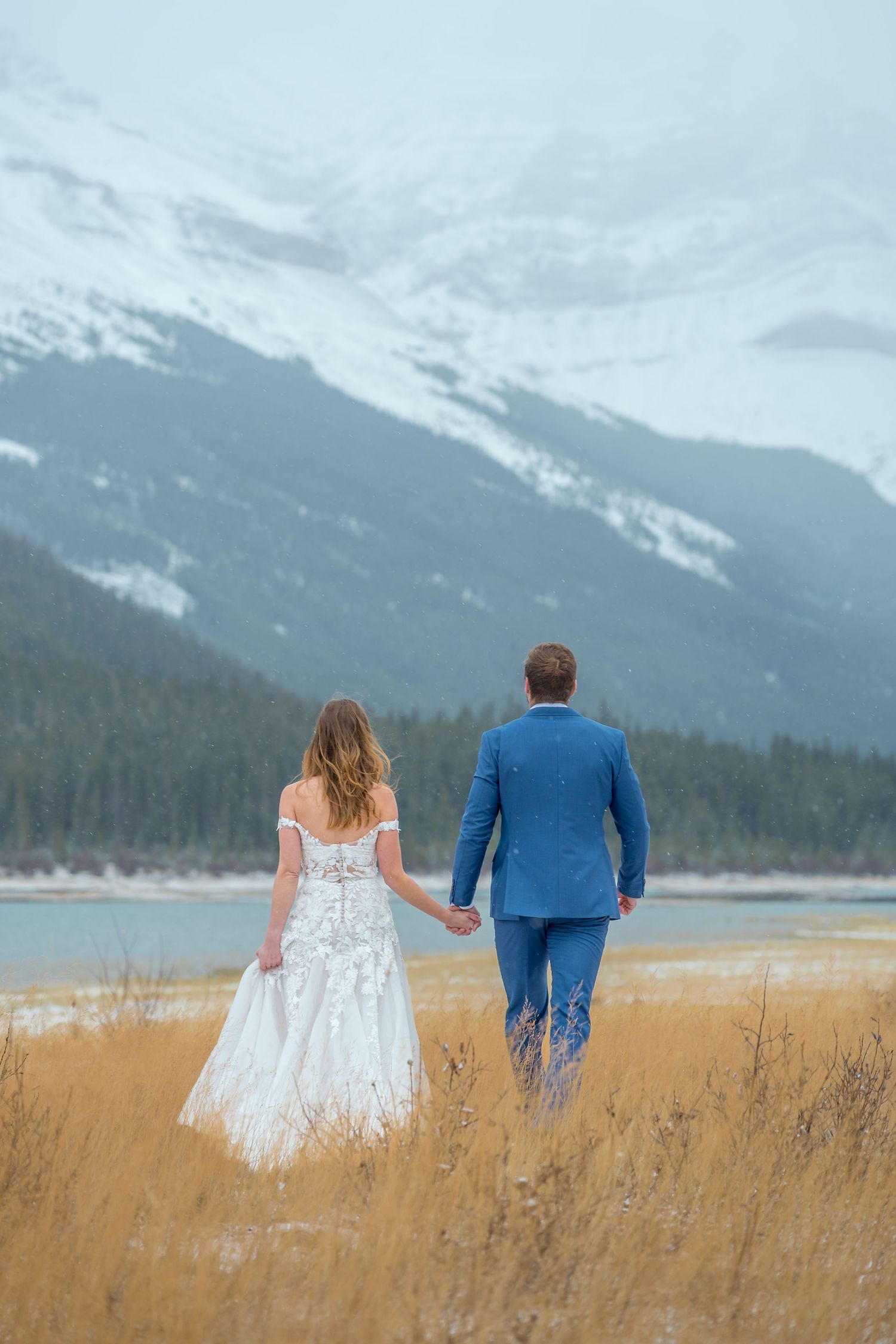 Bride and groom holding hands walking through golden fields with snow-capped mountains and lake backdrop.