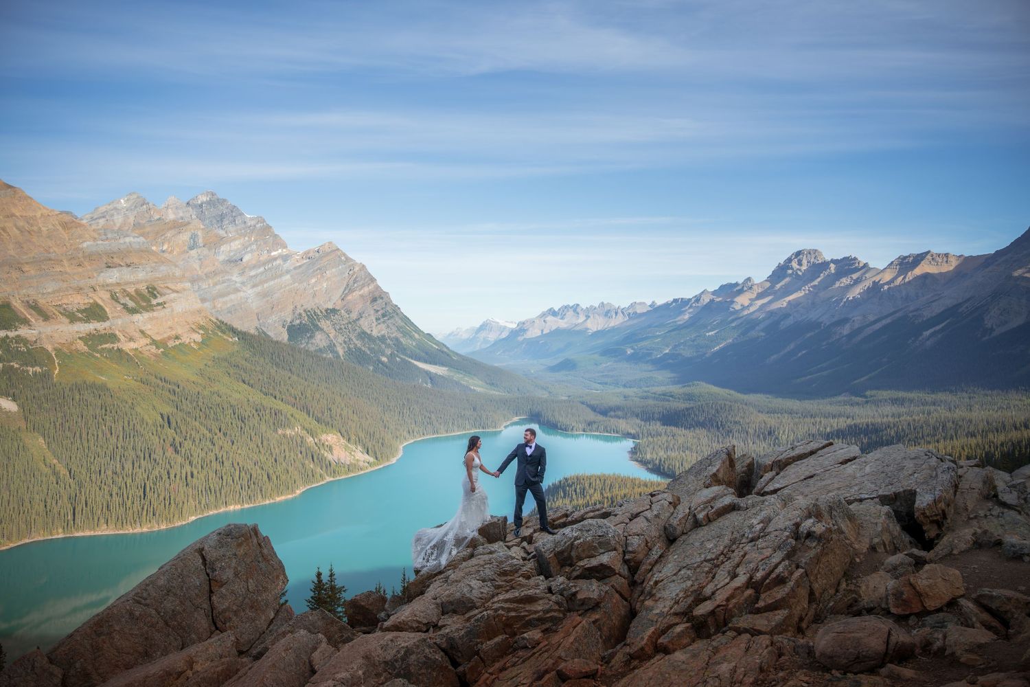 Wedding couple holding hands on rocky overlook above turquoise Peyto Lake surrounded by Canadian Rockies mountains.