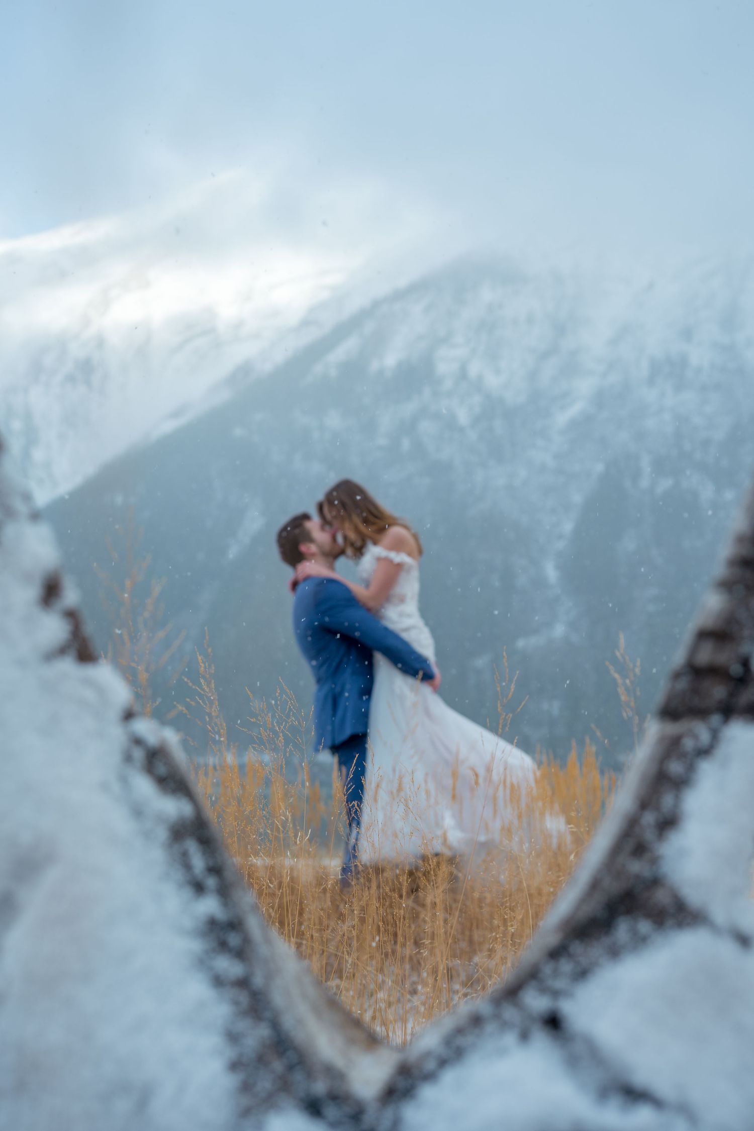 Couple sharing a romantic kiss in snowy mountain landscape, framed by a heart-shaped rock opening on their winter wedding day.